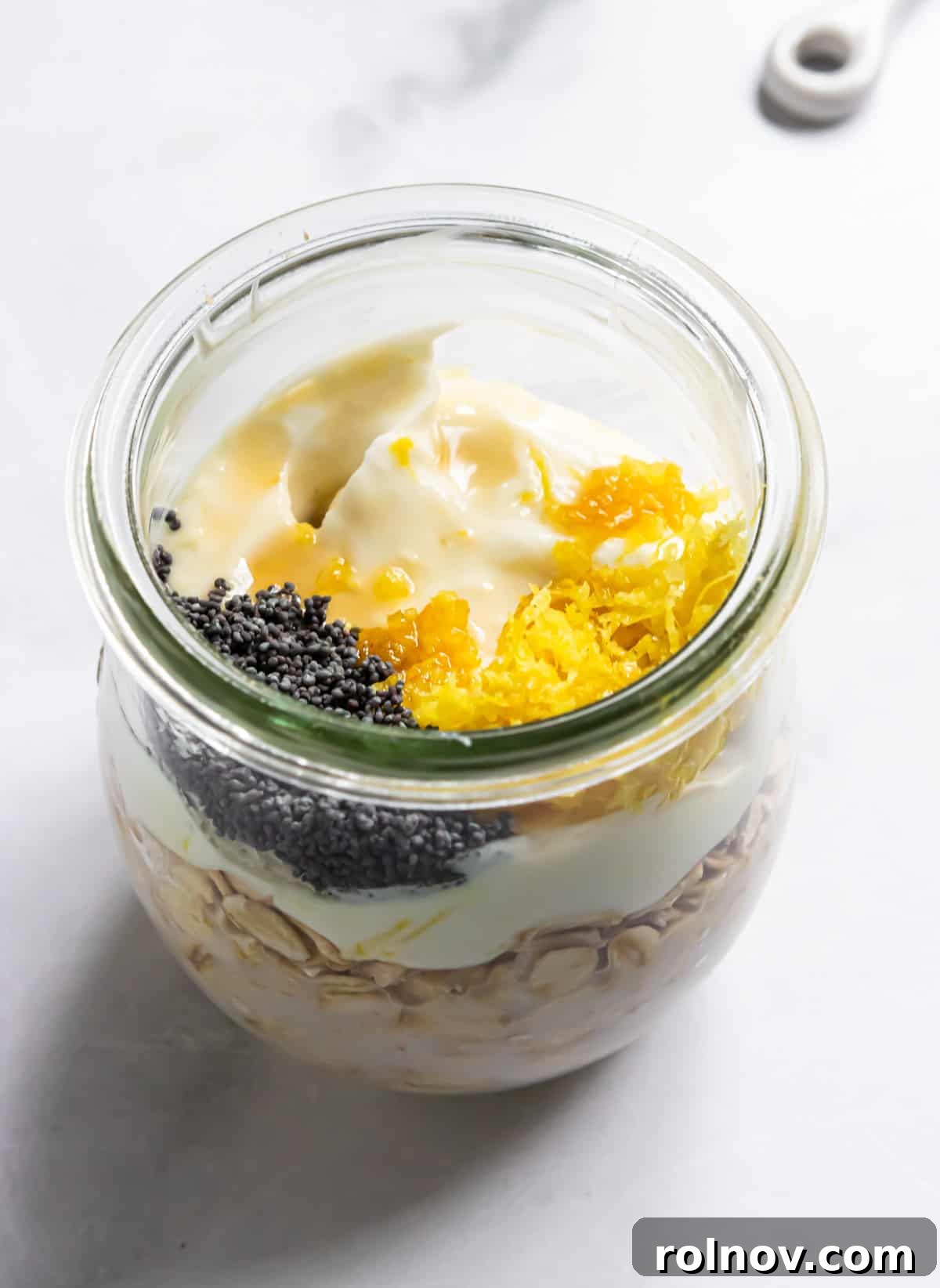 Sunshine Lemon Poppy Seed Overnight Oats 5 Close-up of a jar with mixed ingredients for overnight oats, waiting to be chilled. Visible are oats, milk, and lemon zest.