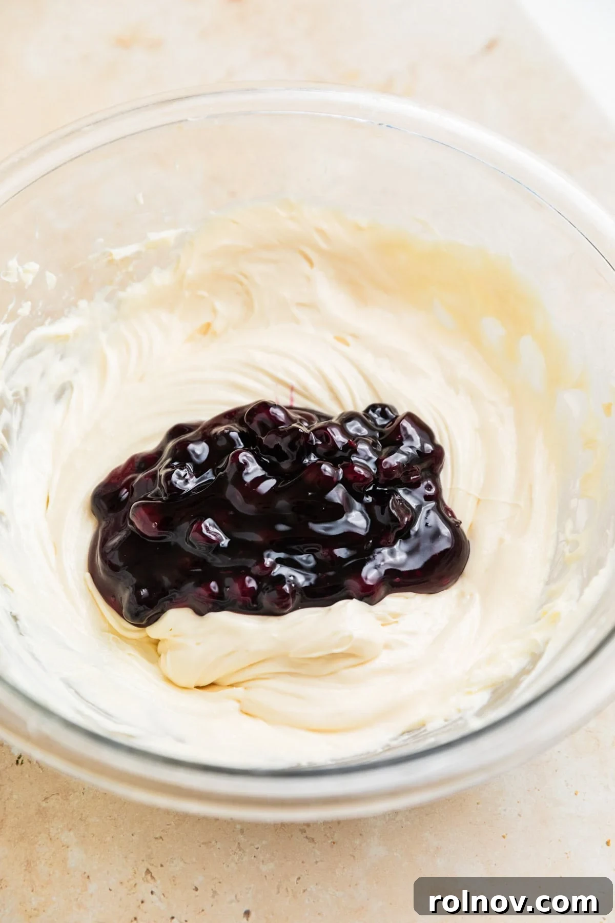 Dreamy Blueberry Cheesecake Dip 6 One cup of vibrant blueberry pie filling gently folded into the smooth cheesecake dip mixture in a clear glass bowl, creating swirls.