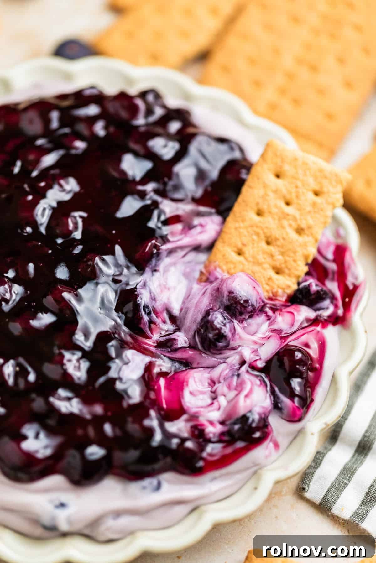 Dreamy Blueberry Cheesecake Dip 8 Close-up of a luscious blueberry cheesecake dip, crowned with glistening blueberry pie filling, with a crispy graham cracker poised for dipping.