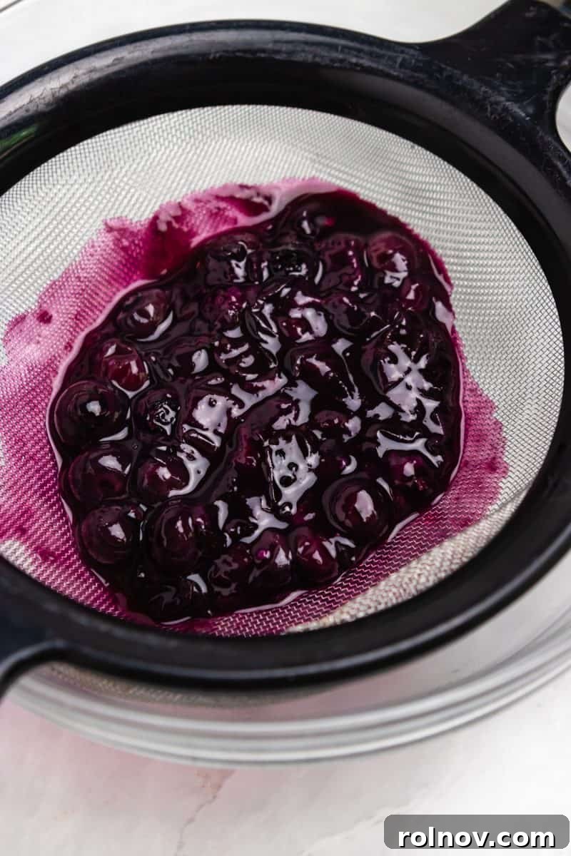 Sweet & Savory Blueberry Basil Goat Cheese Grill 4 Close-up of a small metal strainer holding cooked and mashed blueberries over a bowl, illustrating the straining process for the blueberry sauce.