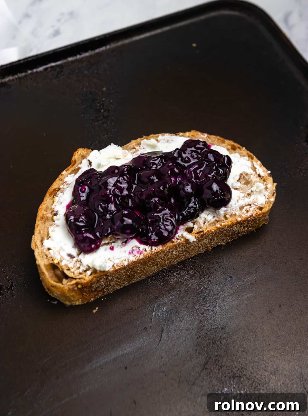 Sweet & Savory Blueberry Basil Goat Cheese Grill 5 Two slices of bread in a hot skillet, with goat cheese spread on both. One slice has blueberry sauce and fresh basil leaves on top, ready to be assembled into a sandwich.