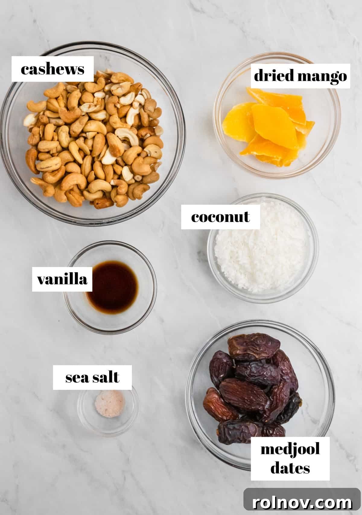 Raw cashews, dried mango, Medjool dates, shredded coconut, and vanilla extract laid out on a kitchen counter, ready for the recipe.