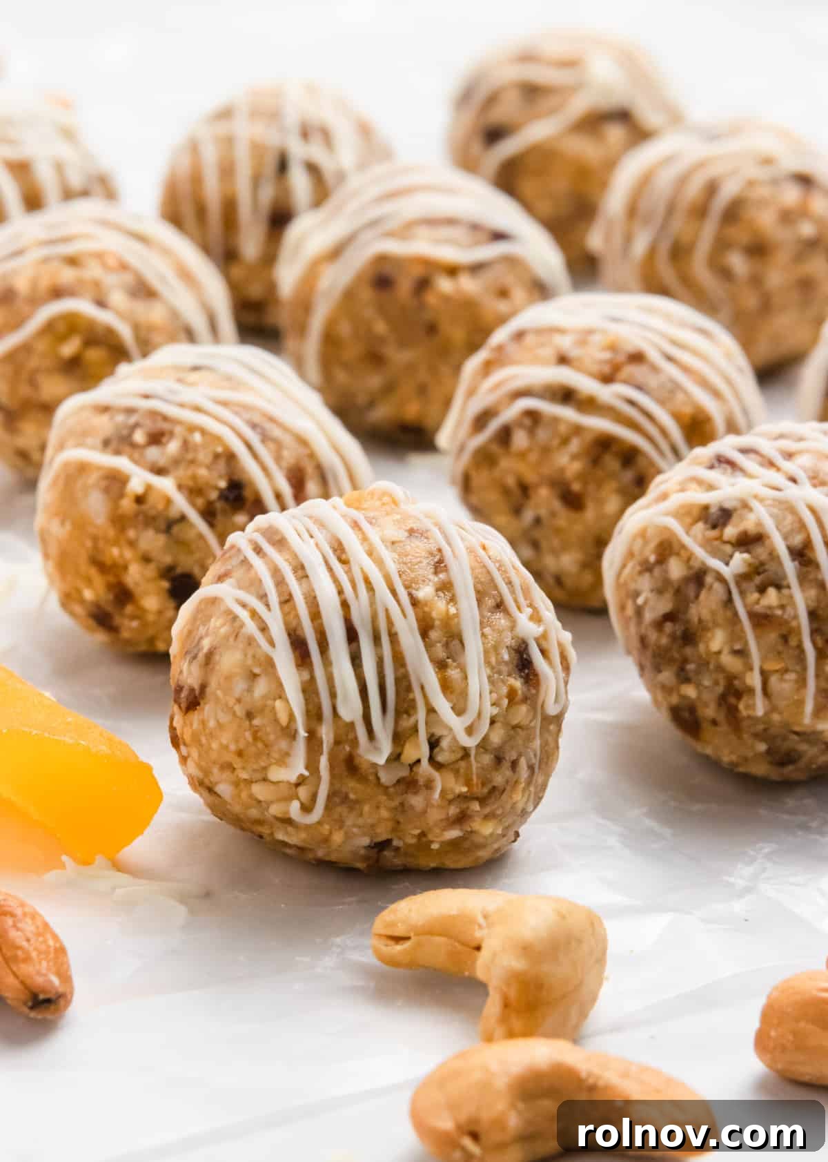 Finished mango energy bites on parchment paper, some drizzled with white chocolate, alongside whole cashews.