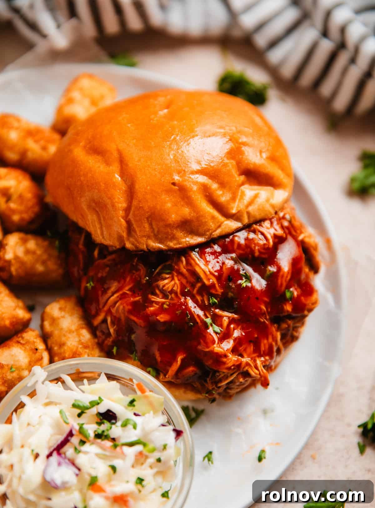 Effortless Slow Cooker BBQ Pulled Chicken 2 Crock pot shredded BBQ chicken served on a brioche bun with extra BBQ sauce, a side of coleslaw, and crispy tater tots.