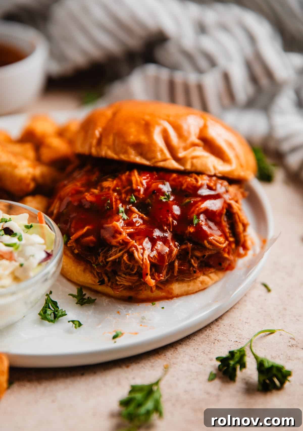 Effortless Slow Cooker BBQ Pulled Chicken 8 A delicious pulled BBQ chicken sandwich on a white plate, accompanied by a fresh side of coleslaw and crispy tater tots.