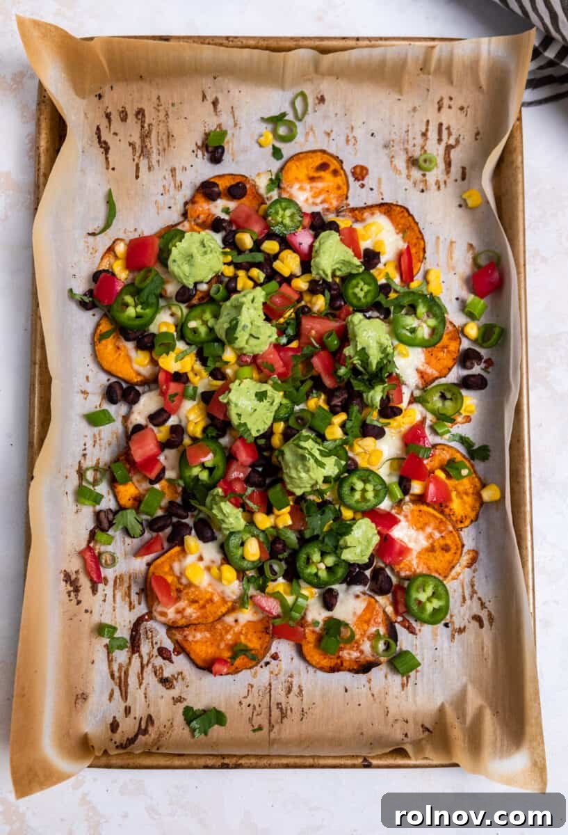Overhead view of a sheet pan laden with fully loaded sweet potato nachos, garnished with avocado crema, tomato, and fresh herbs.