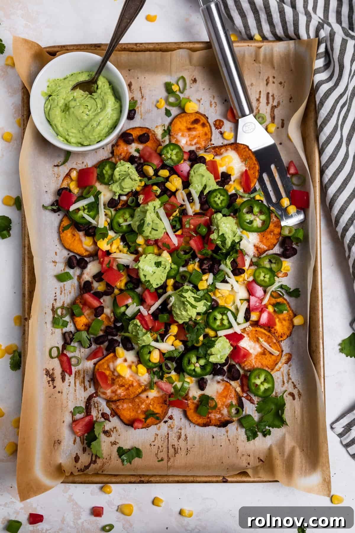 Overhead view of a sheet pan filled with loaded sweet potato nachos alongside a bowl of creamy avocado crema.