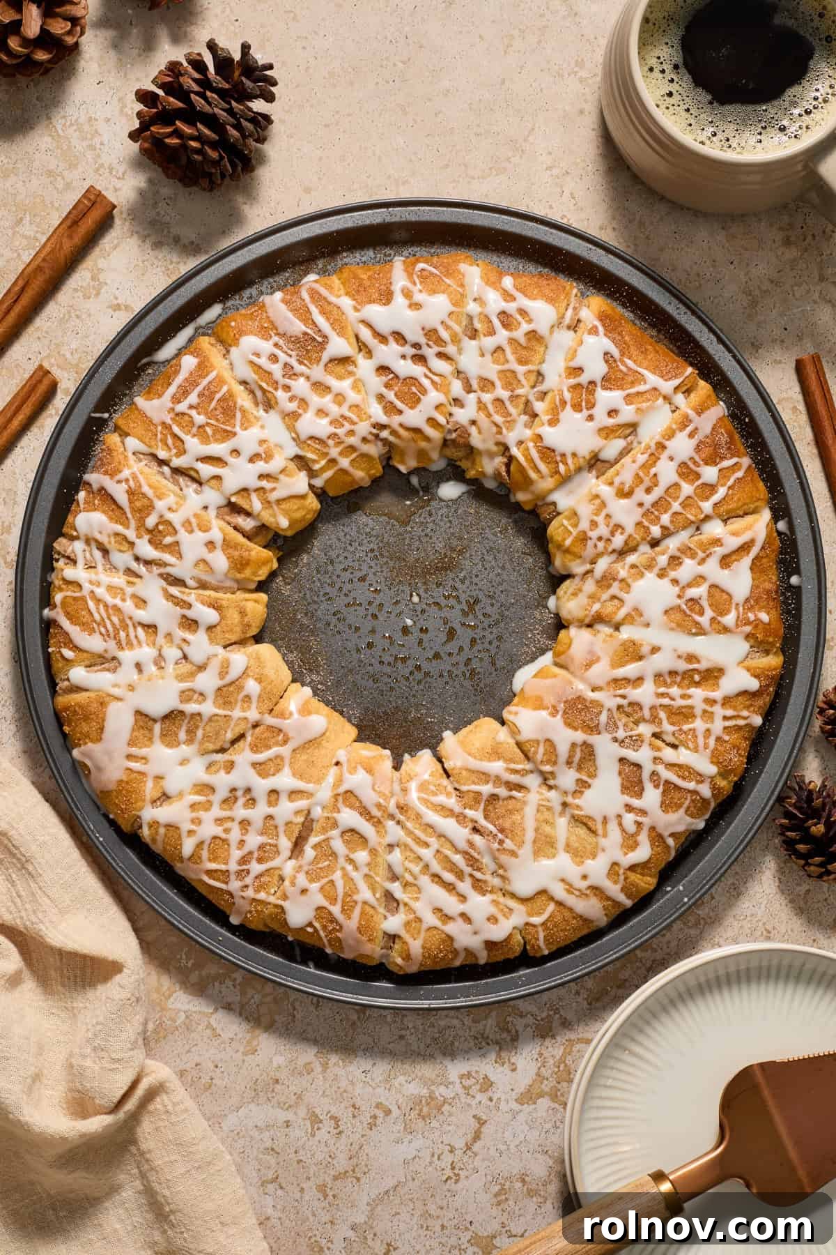 A festive cinnamon cream cheese crescent roll wreath on a baking pan, garnished with cinnamon sticks and a cup of coffee nearby, ready for a holiday breakfast.