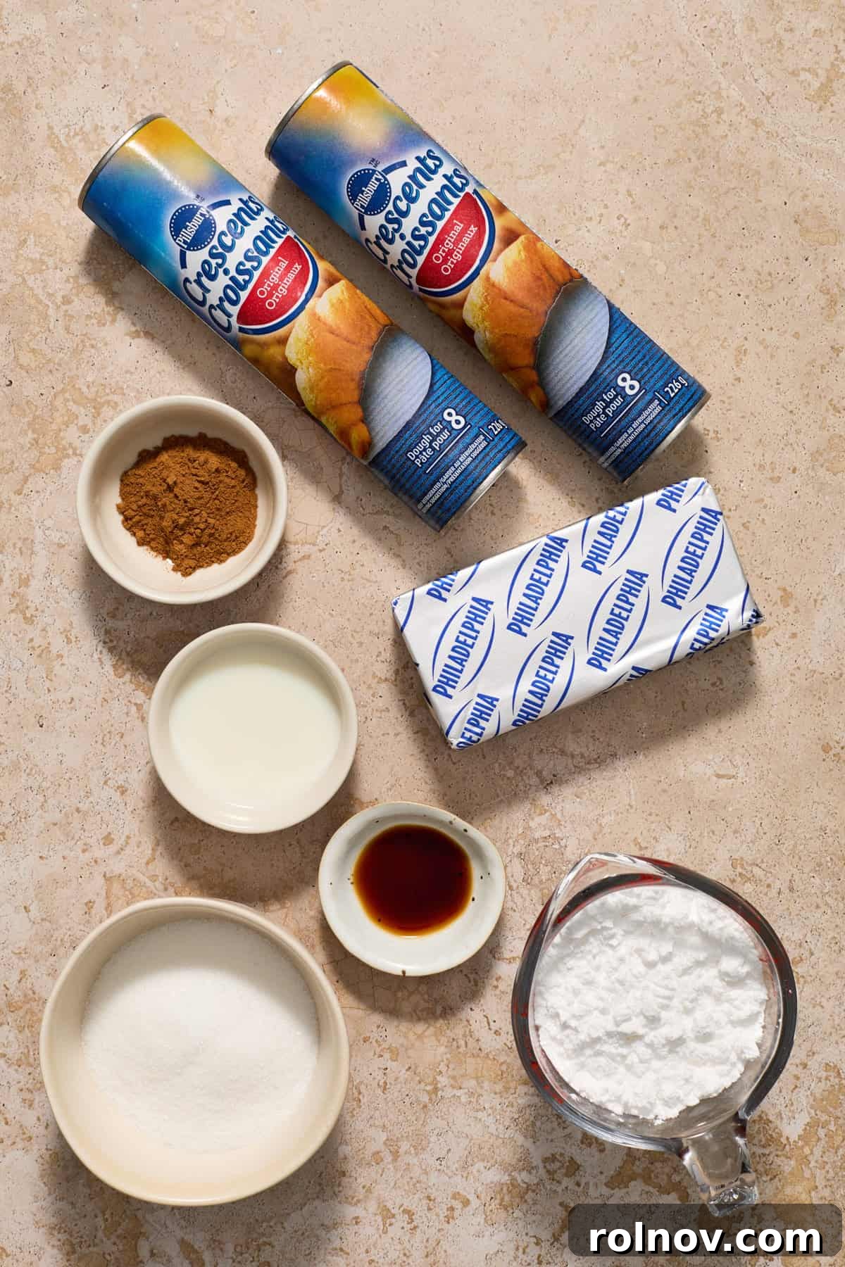 A collection of ingredients for the crescent roll wreath, including crescent dough, powdered sugar, cream cheese, and spices, laid out on a kitchen surface.