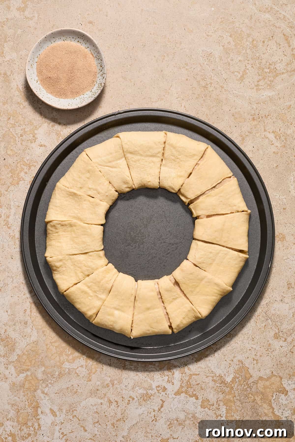 Crescent roll wreath on cookie sheet before baking with cinnamon and sugar in bowl beside it.