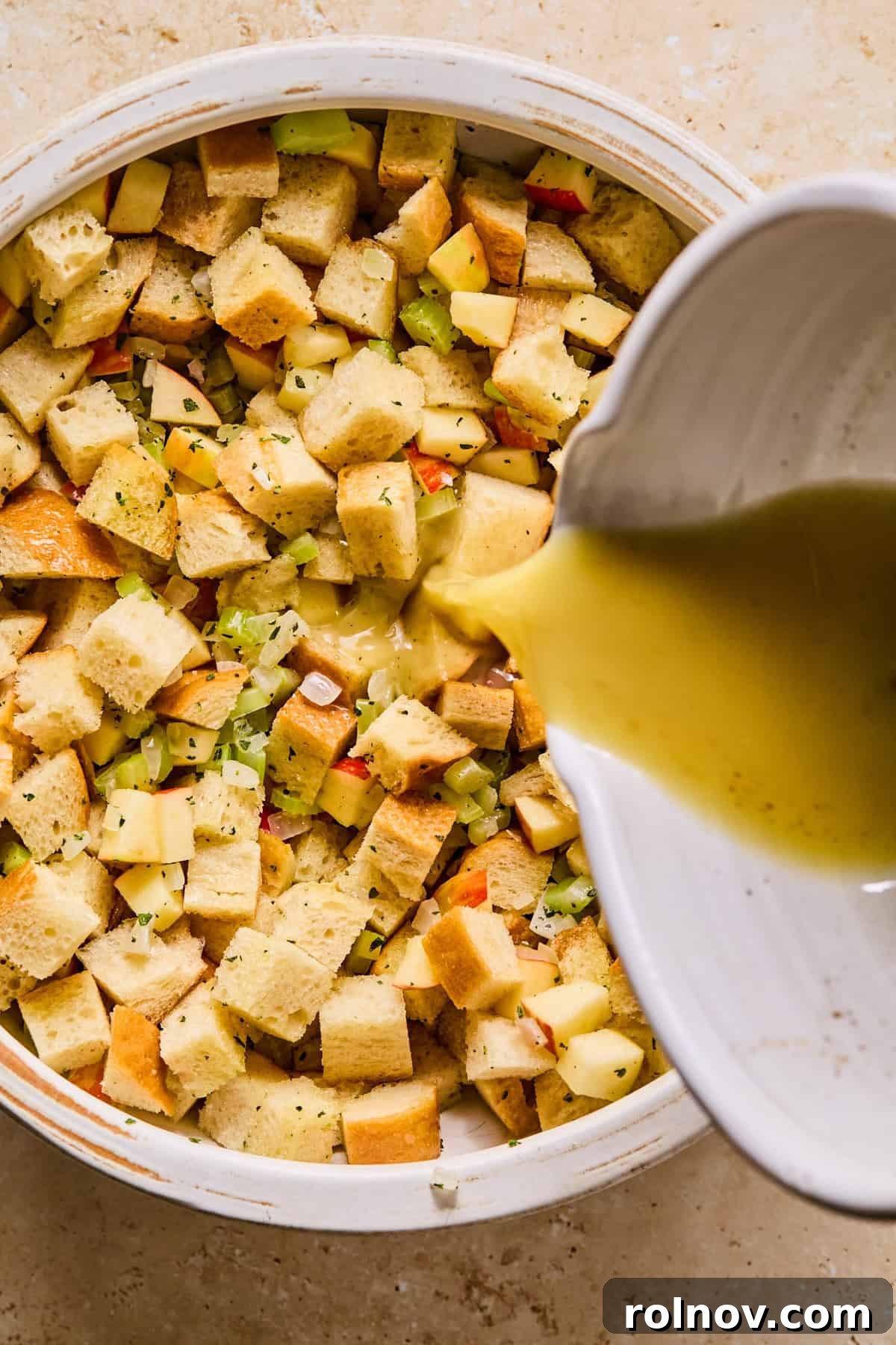 Homestyle Apple Sage Stuffing 9 Liquid mixture being poured over the bread, apple, celery, and onion in a large bowl.