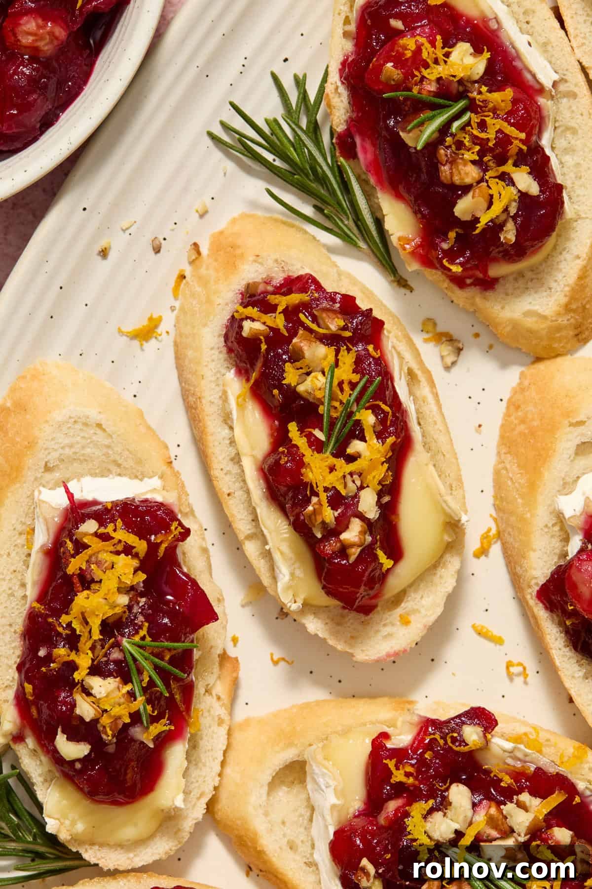 Close-up of a Cranberry Brie Crostini on a plate, topped with fresh rosemary and orange zest, showcasing its delightful textures.