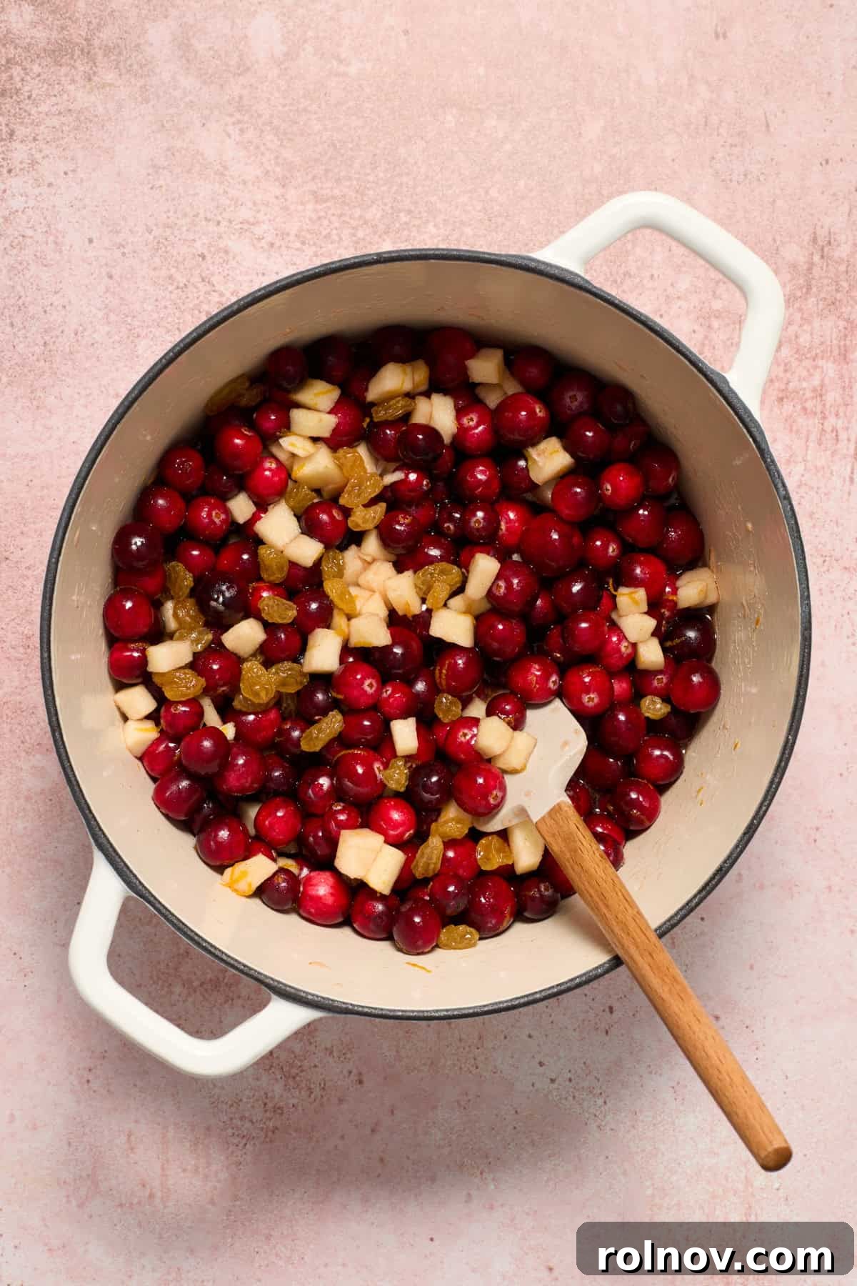 Pot with fresh cranberries, diced apples, golden raisins, and a spatula, ready to be cooked into chutney.