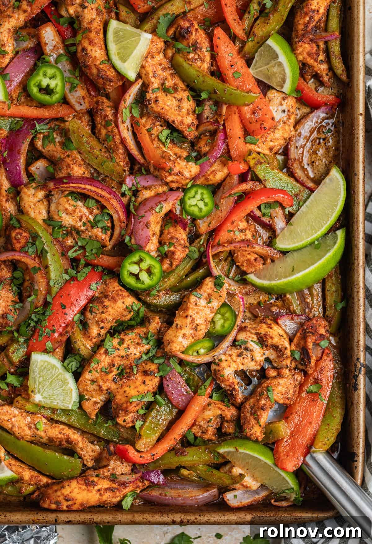Baked chicken fajitas on sheet pan with onions and peppers, topped with chopped cilantro, lime wedges and jalapeños.