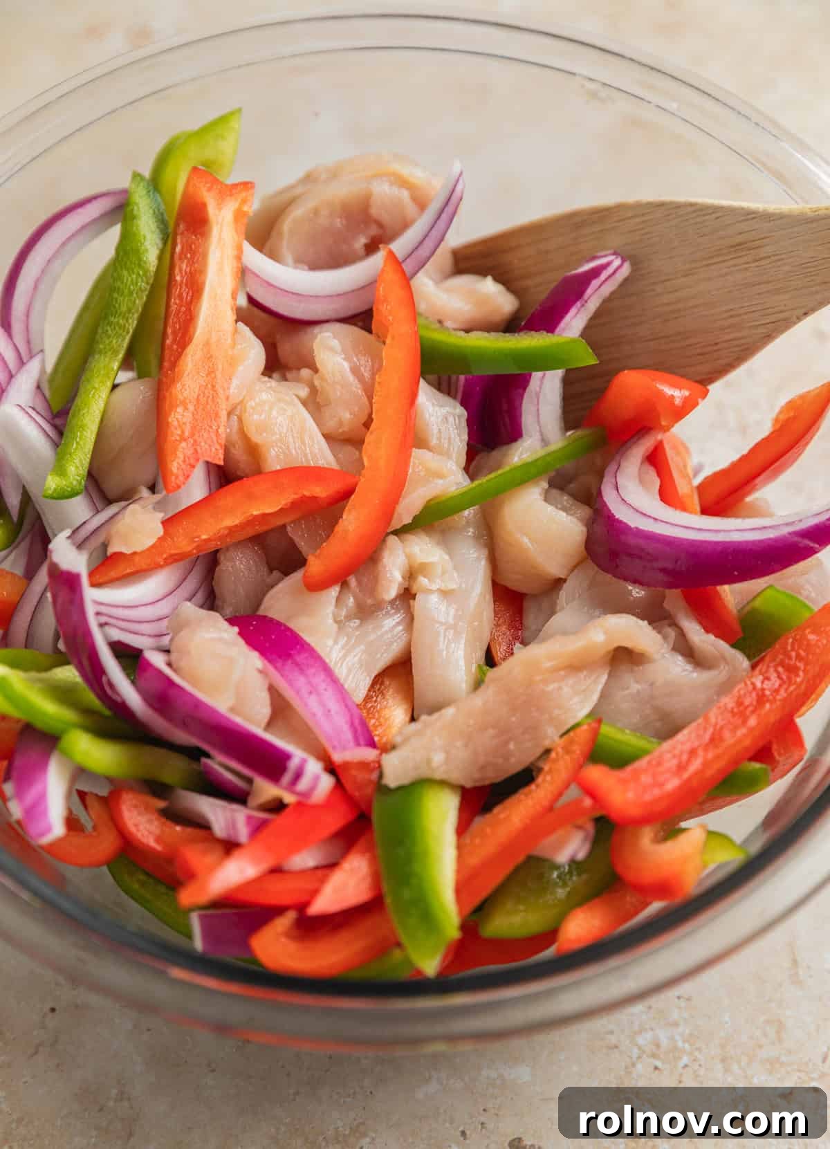 Raw sliced chicken, sliced red and green bell peppers and red onion in glass bowl with wood spoon.