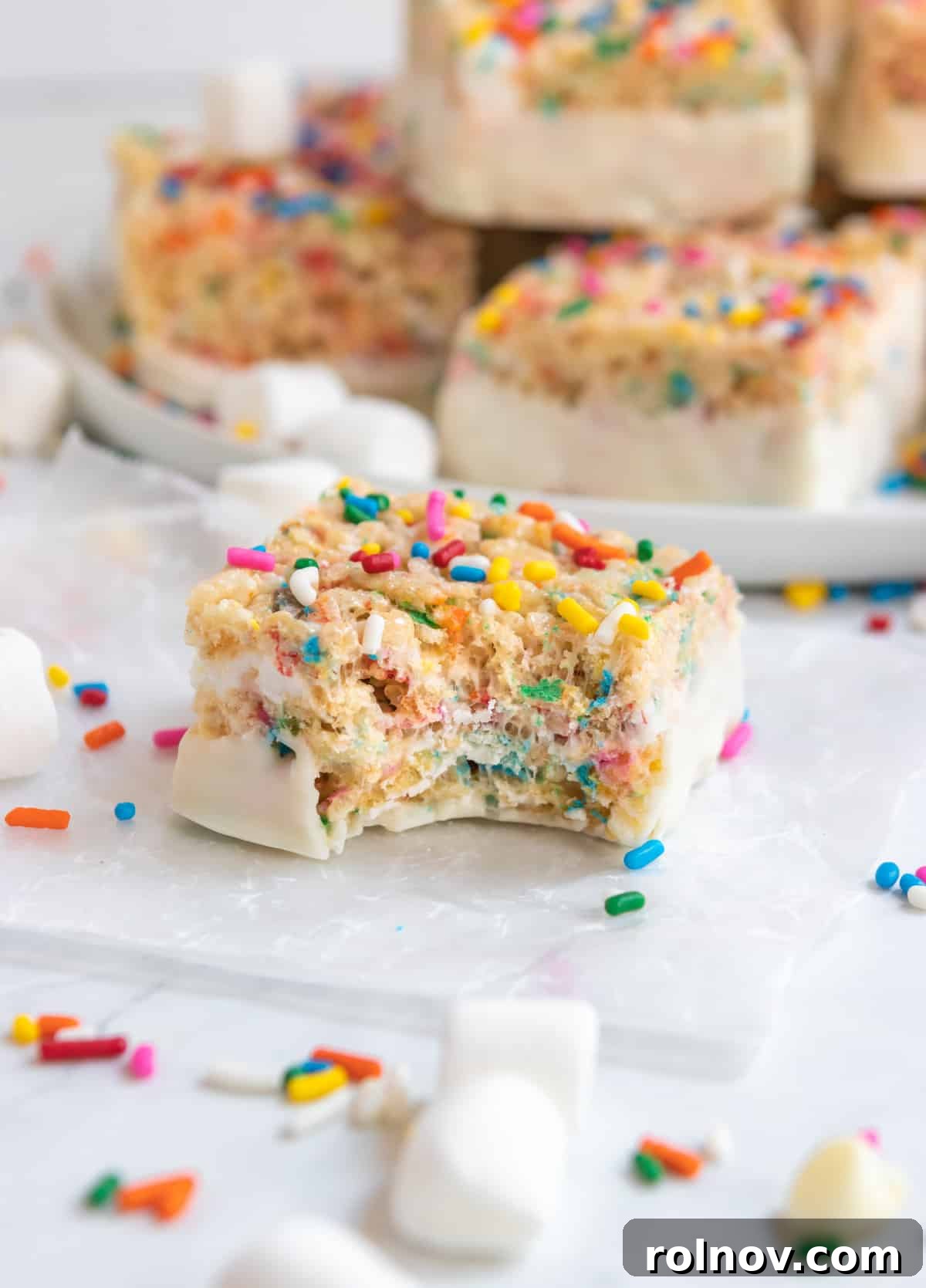 Festive Sprinkled Krispie Treats 2 Vibrant Funfetti Rice Krispie Treats with rainbow sprinkles, partially dipped in white chocolate and resting on parchment paper.