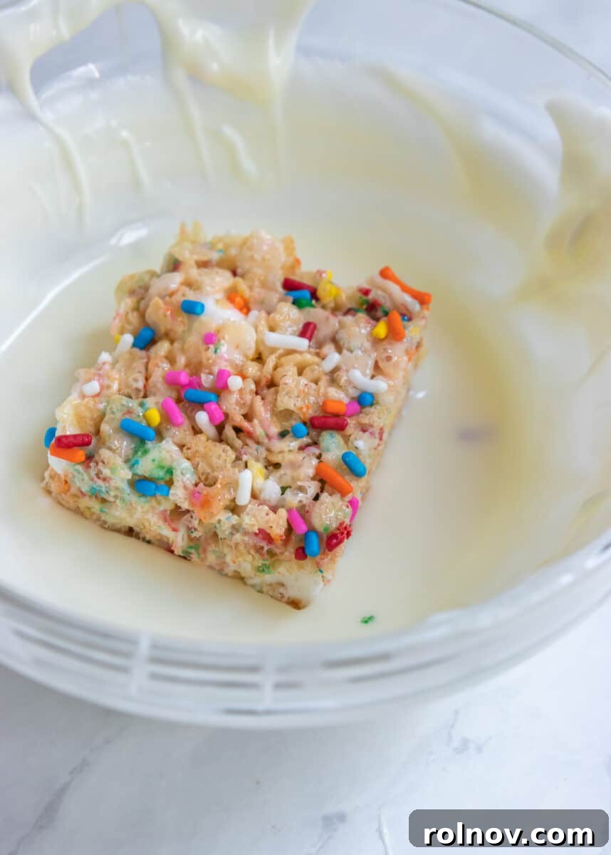 Festive Sprinkled Krispie Treats 7 A single Funfetti Rice Krispie Treat being dipped into a bowl of melted white chocolate, showing the smooth coating process.