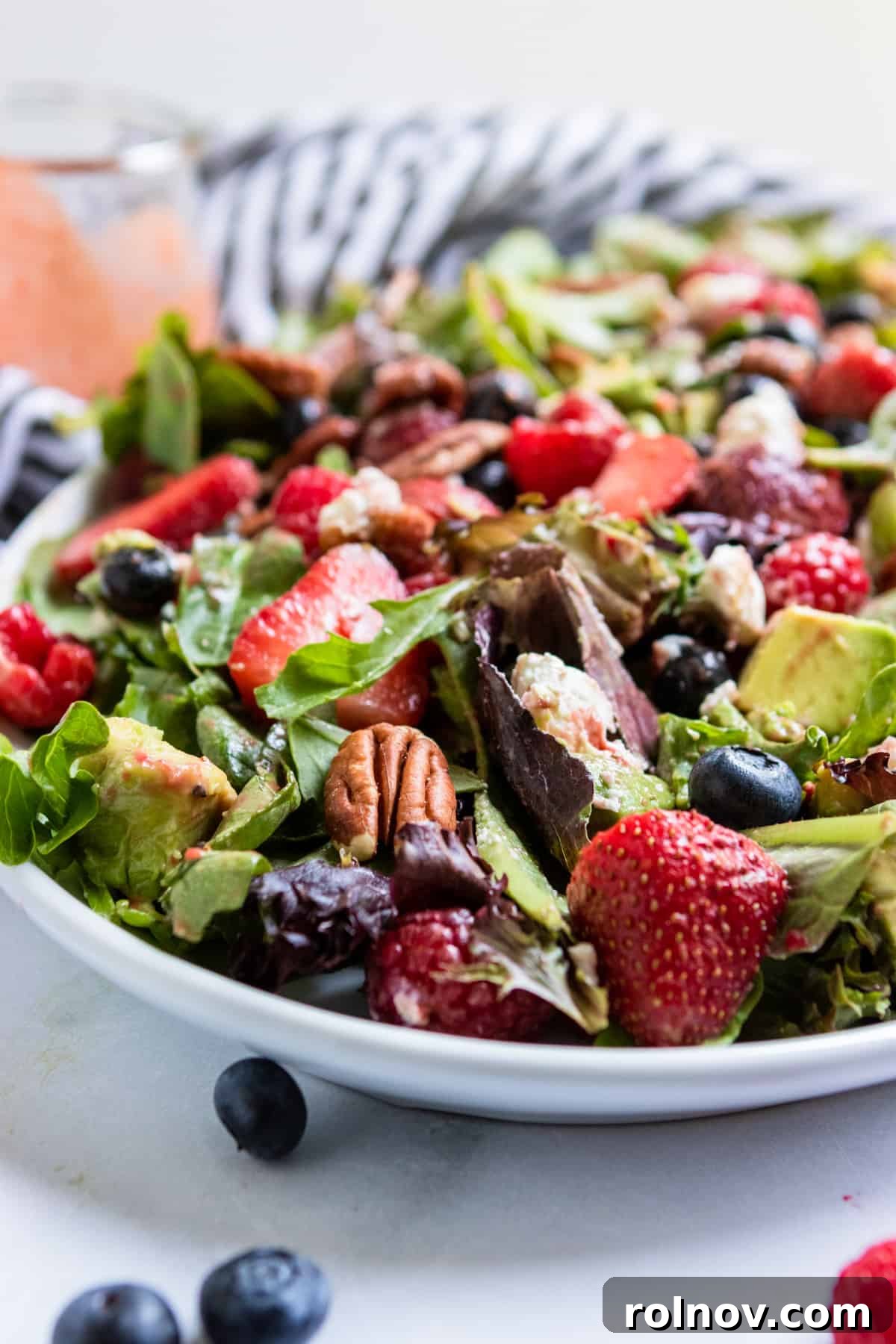 Strawberry Balsamic Berry Salad 2 Mixed green salad with assorted berries and goat cheese on a white plate, with a small bowl of strawberry balsamic dressing on the side.