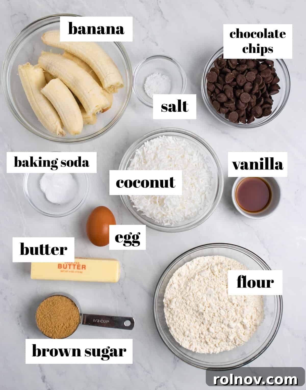 Individual ingredients like flour, chocolate chips, banana, butter, and an egg are labeled on a kitchen counter, ready for baking.