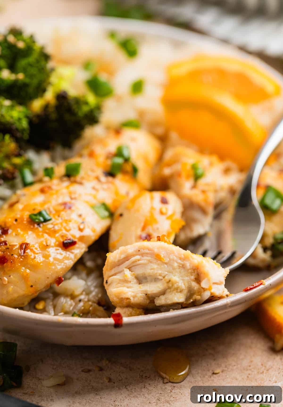 Sticky Orange Chicken One-Pan Dinner 3 Piece of baked orange chicken on fork with plate of broccoli and rice.
