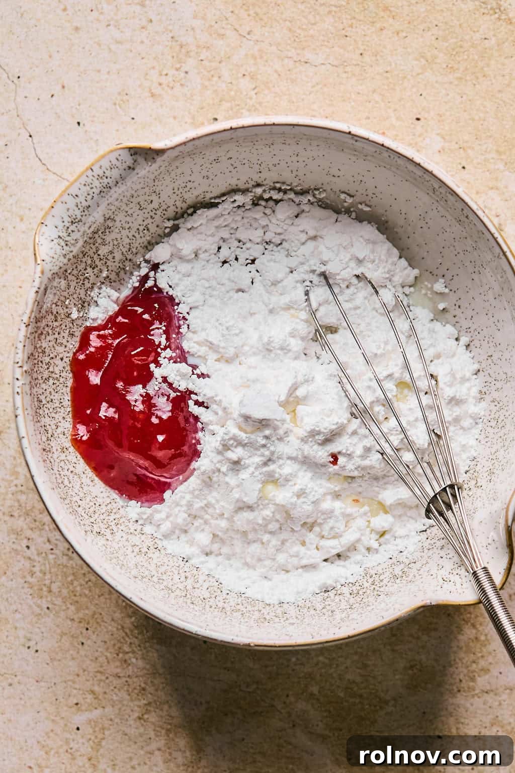 A bowl containing strawberry jam, powdered sugar, and other icing ingredients, alongside a whisk, ready for mixing.