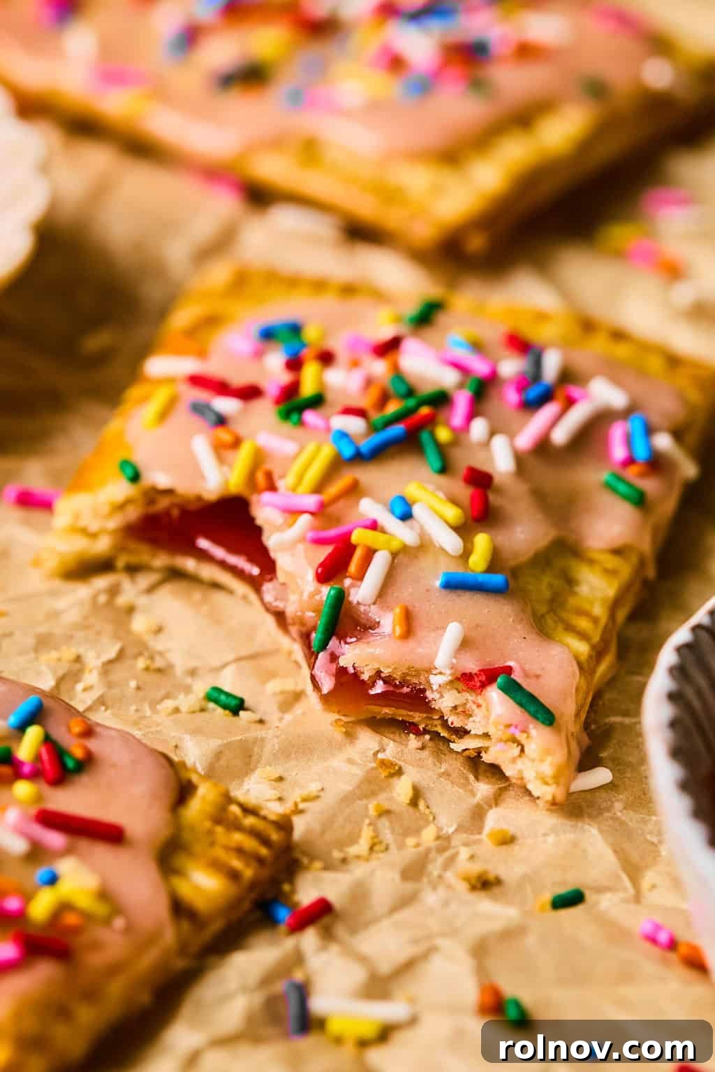 A baked homemade pop tart filled with strawberry jam, with the jam slightly oozing, on parchment paper with rainbow sprinkles.