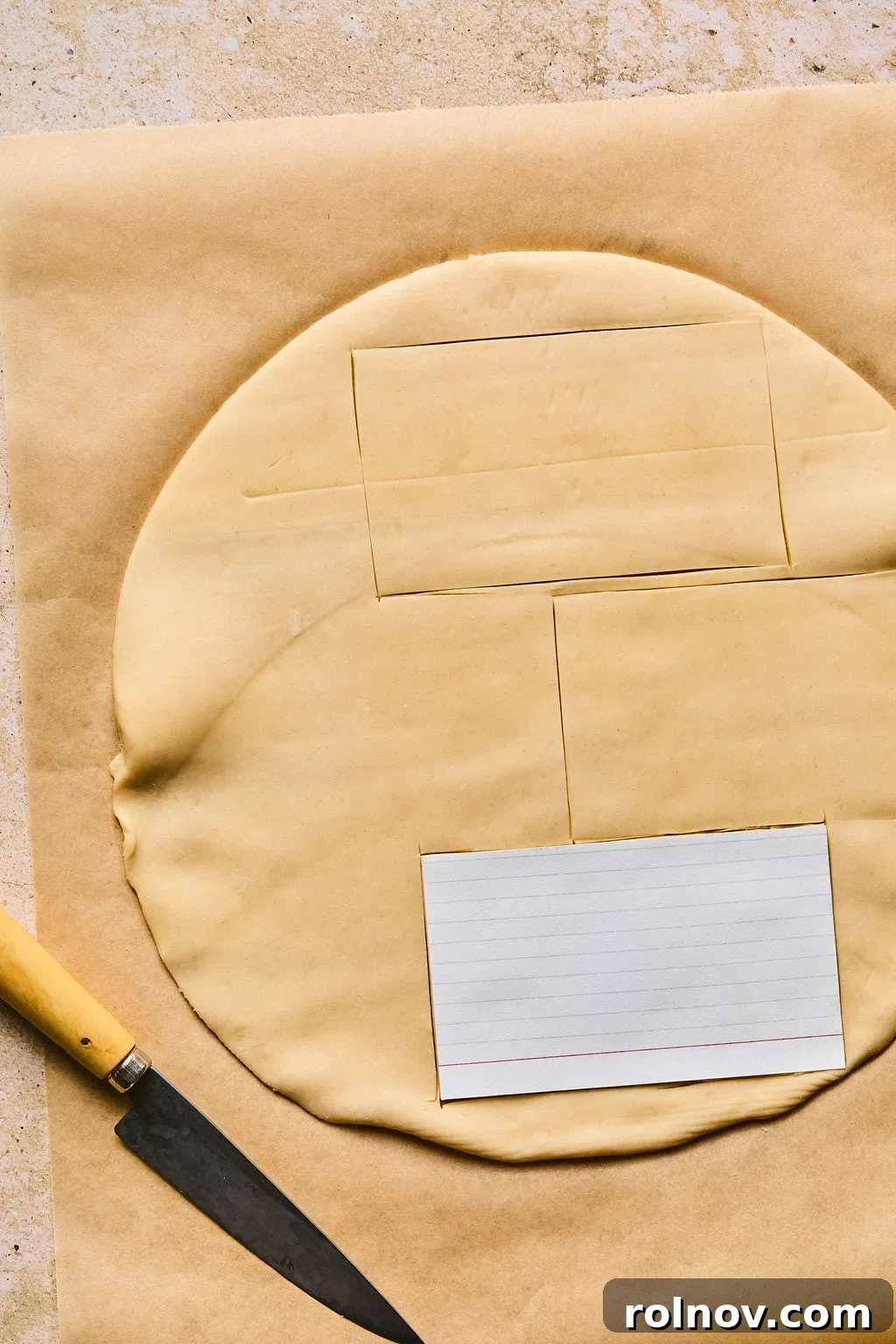 Unrolled pie crust on a cutting board with an index card used as a stencil to guide cutting rectangular shapes.