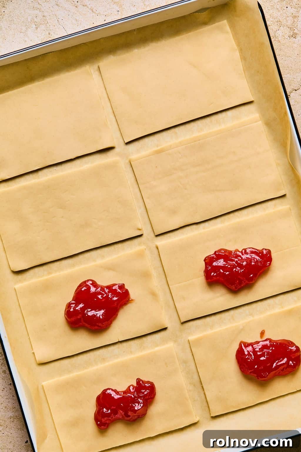 Small dollops of bright strawberry jam placed precisely in the center of individual pie crust rectangles on a baking sheet.