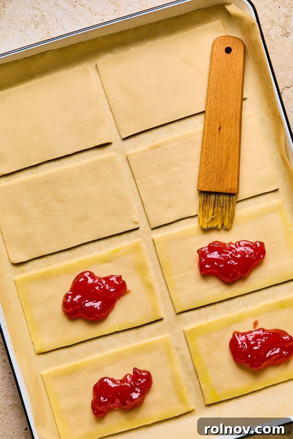 A pastry brush applies an even layer of egg wash along the perimeter of a pie crust rectangle, surrounding the jam filling.