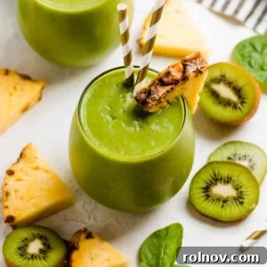 Wine glass with pineapple kiwi smoothie with two straws and sliced pineapple and kiwi surrounding.