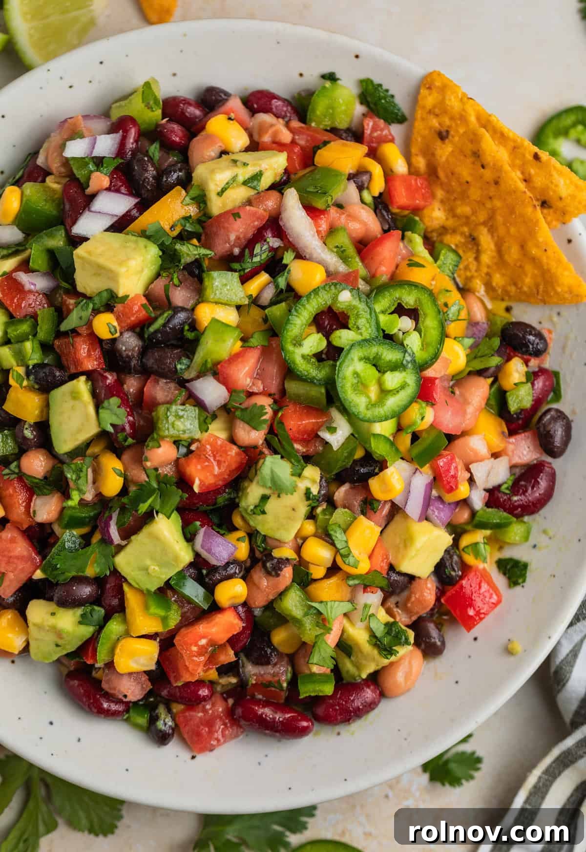 Overhead view of a white bowl filled with a vibrant 3 Bean Avocado Corn Salad, generously topped with fresh chopped cilantro. The salad features a colorful mix of beans, corn, bell peppers, and avocado.