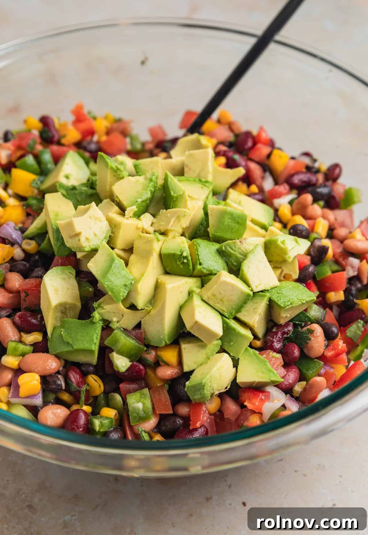 A close-up of the partially mixed bean, corn, and vegetable salad in a large bowl, showcasing its vibrant colors before the addition of avocado, with a spoon resting in the bowl.