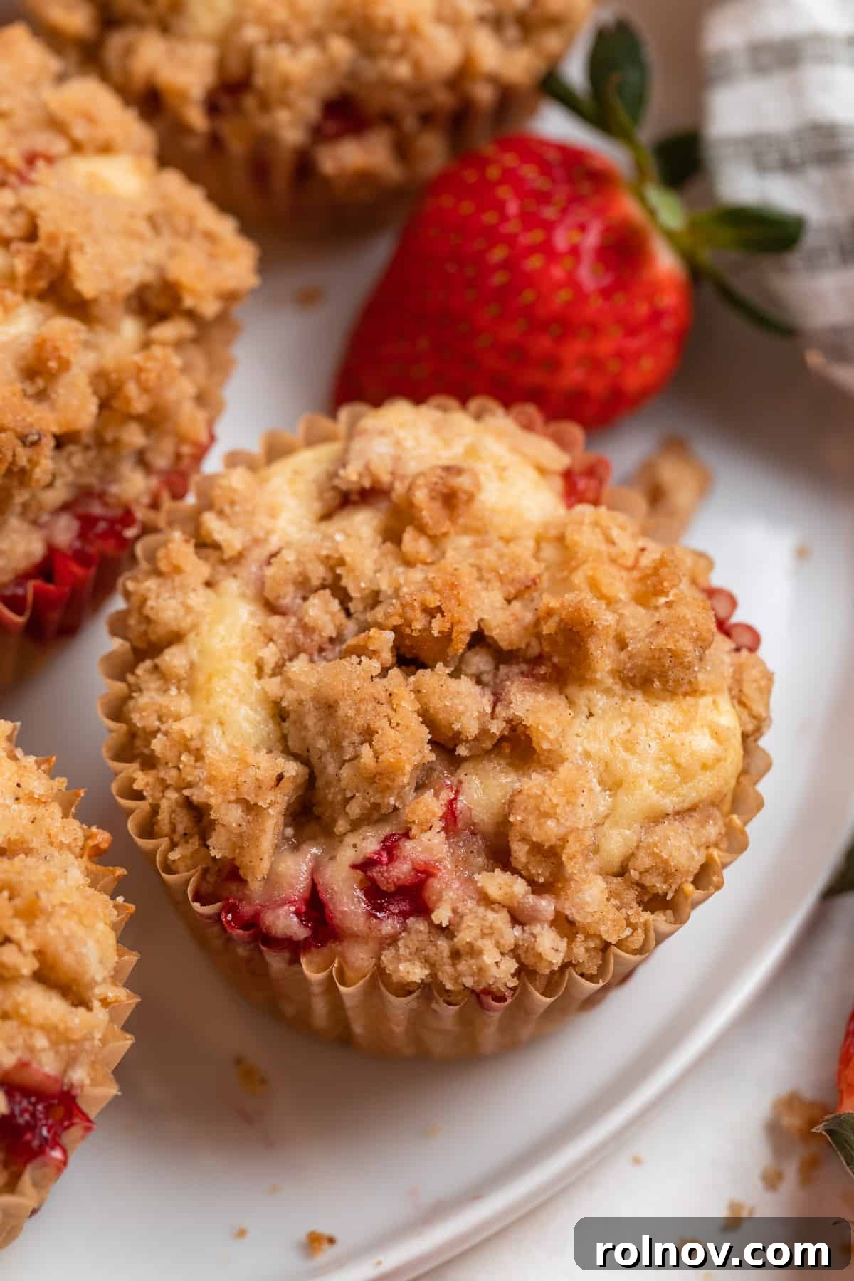 Sweet Strawberry Crumble Muffins 2 Freshly baked strawberry crumble muffins on a white plate, surrounded by whole strawberries.