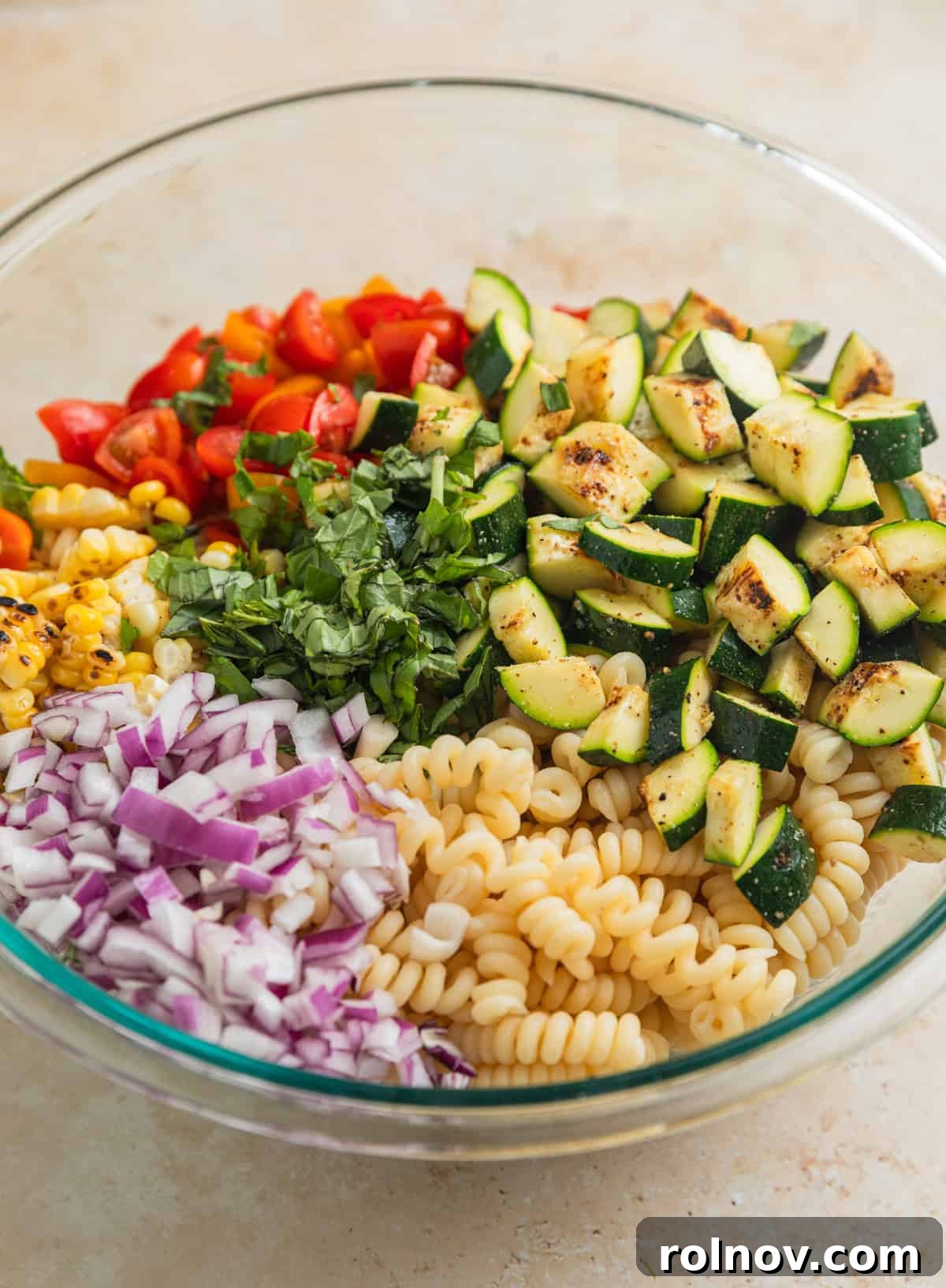 Charred Zucchini Pasta Toss 7 A large glass mixing bowl filled with cooked spiral pasta, chopped red onion, quartered grape tomatoes, grilled corn kernels, and bite-sized grilled zucchini pieces, ready for dressing.
