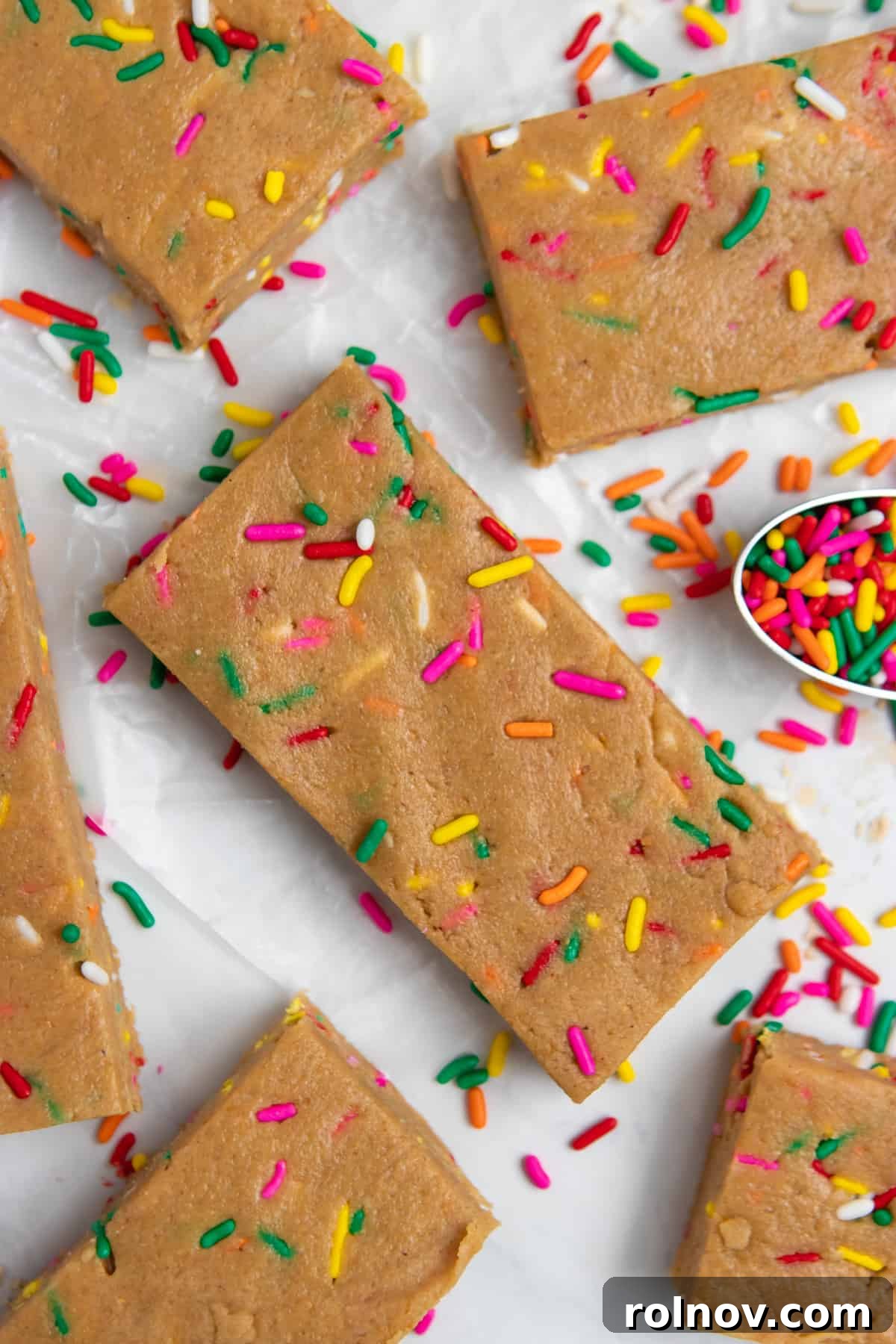 Birthday Cake Protein Bliss 2 No bake birthday cake protein bars on white counter with spoon of sprinkles.