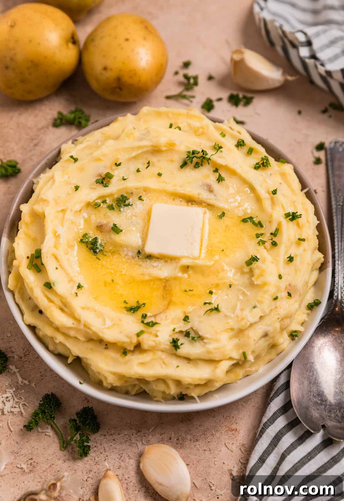 Creamy Instant Pot Garlic Parmesan Mashed Potatoes 3 Garlic Parmesan mashed potatoes in a bowl, surrounded by fresh garlic cloves, parsley, and other ingredients.