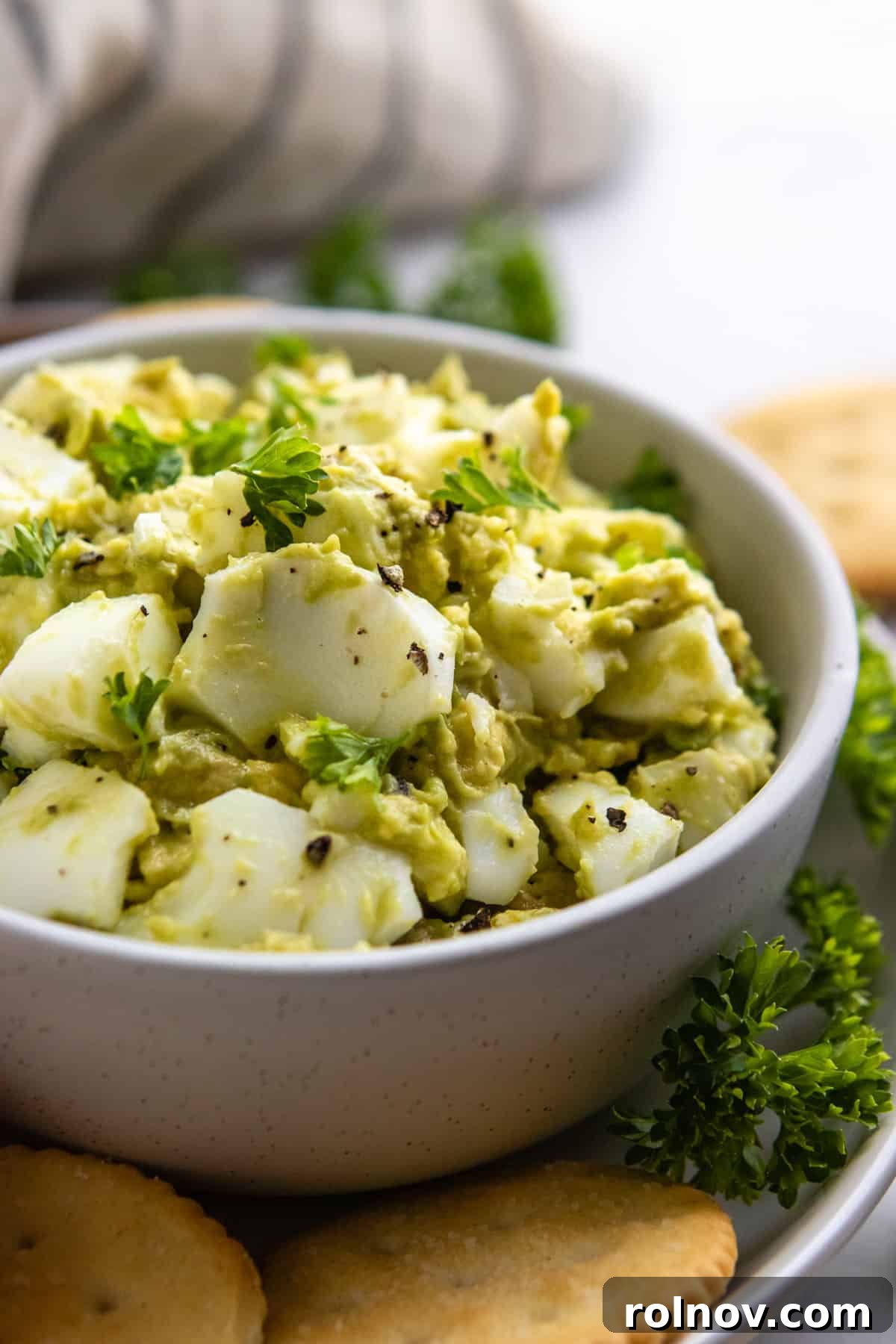 Creamy Avocado Egg Salad 2 Avocado Egg Salad in a rustic bowl, garnished with fresh parsley, surrounded by whole wheat crackers, illustrating a healthy and delicious meal.