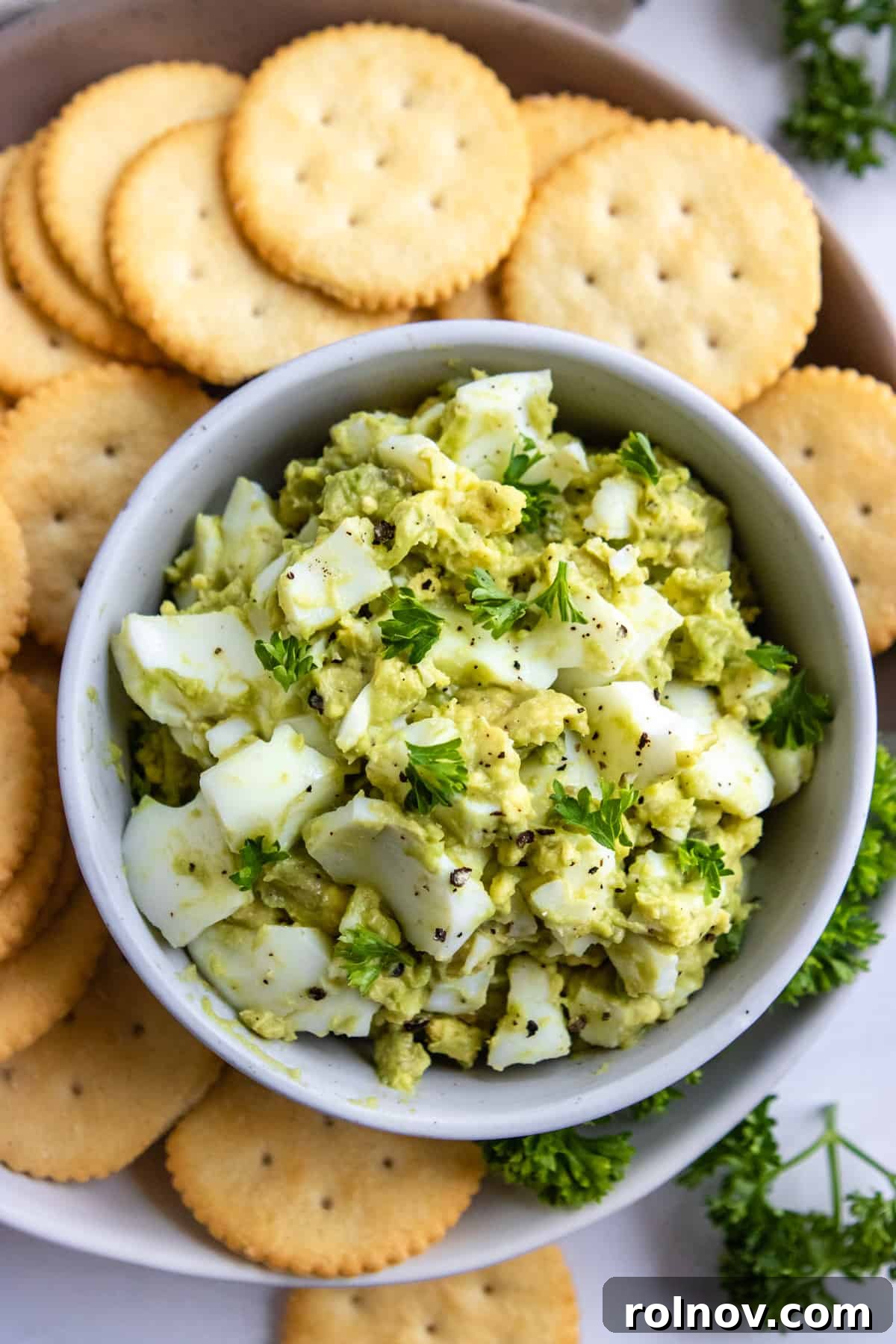 Creamy Avocado Egg Salad 5 An inviting overhead view of a bowl filled with vibrant Avocado Egg Salad, garnished with fresh herbs, accompanied by a side of wholesome crackers for easy dipping.