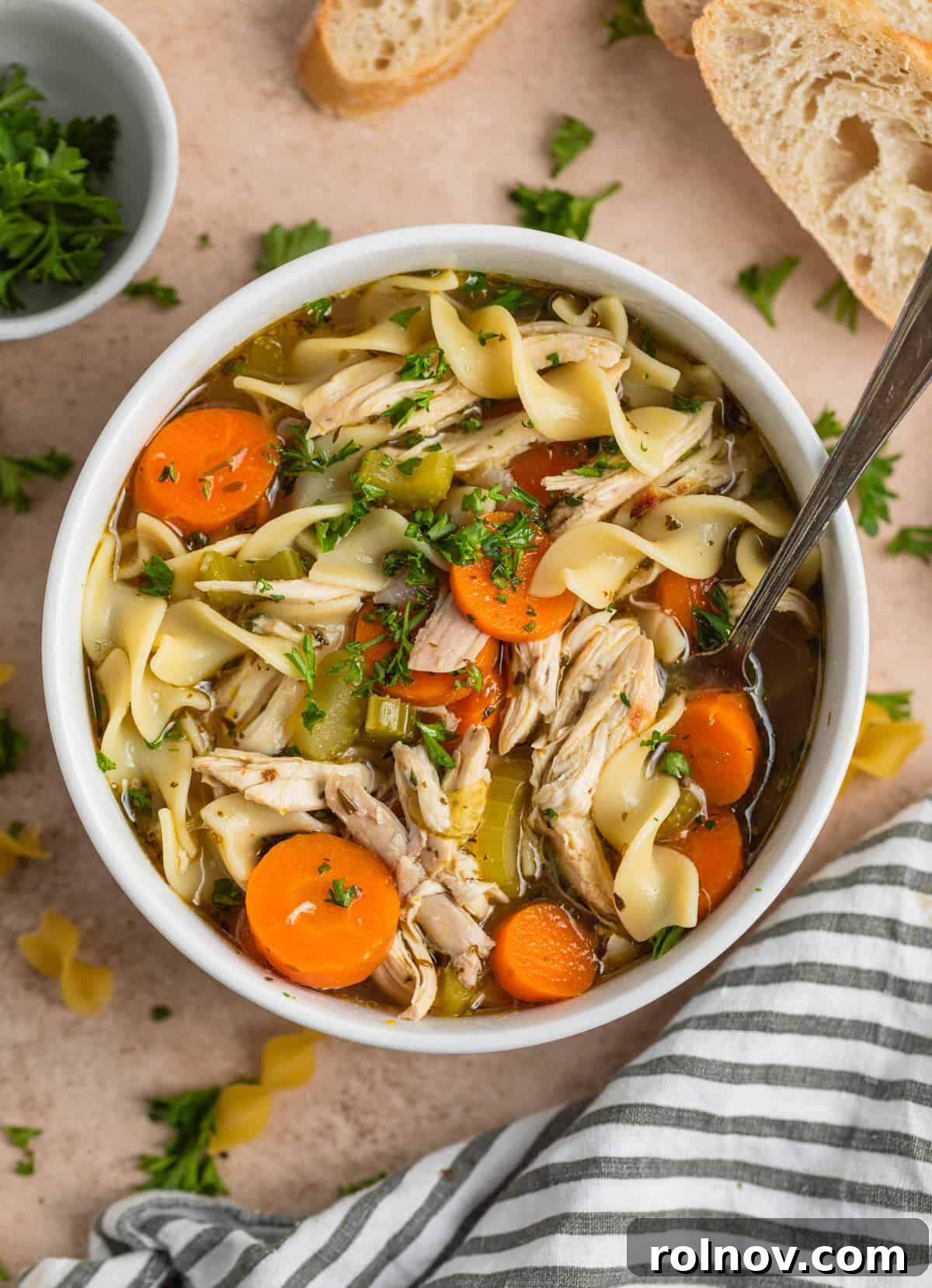 Shortcut Rotisserie Chicken Noodle Soup 2 A steaming bowl of homemade rotisserie chicken noodle soup, garnished with fresh chopped parsley, and a soup spoon ready for enjoyment.