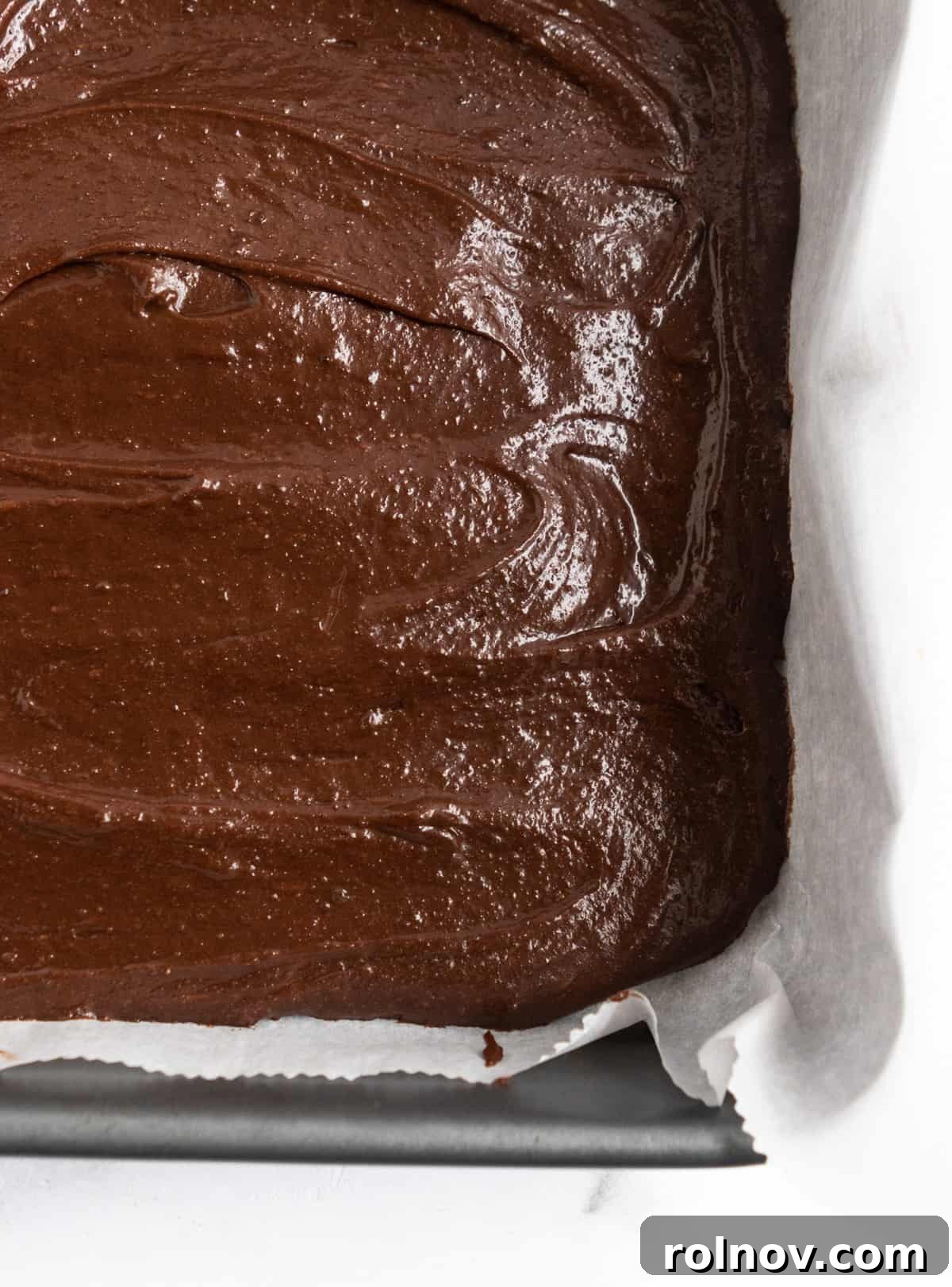 Brownie batter in parchment lined pan. A close-up shot of the spread brownie batter in a parchment-lined square baking pan, ready for the next layer.