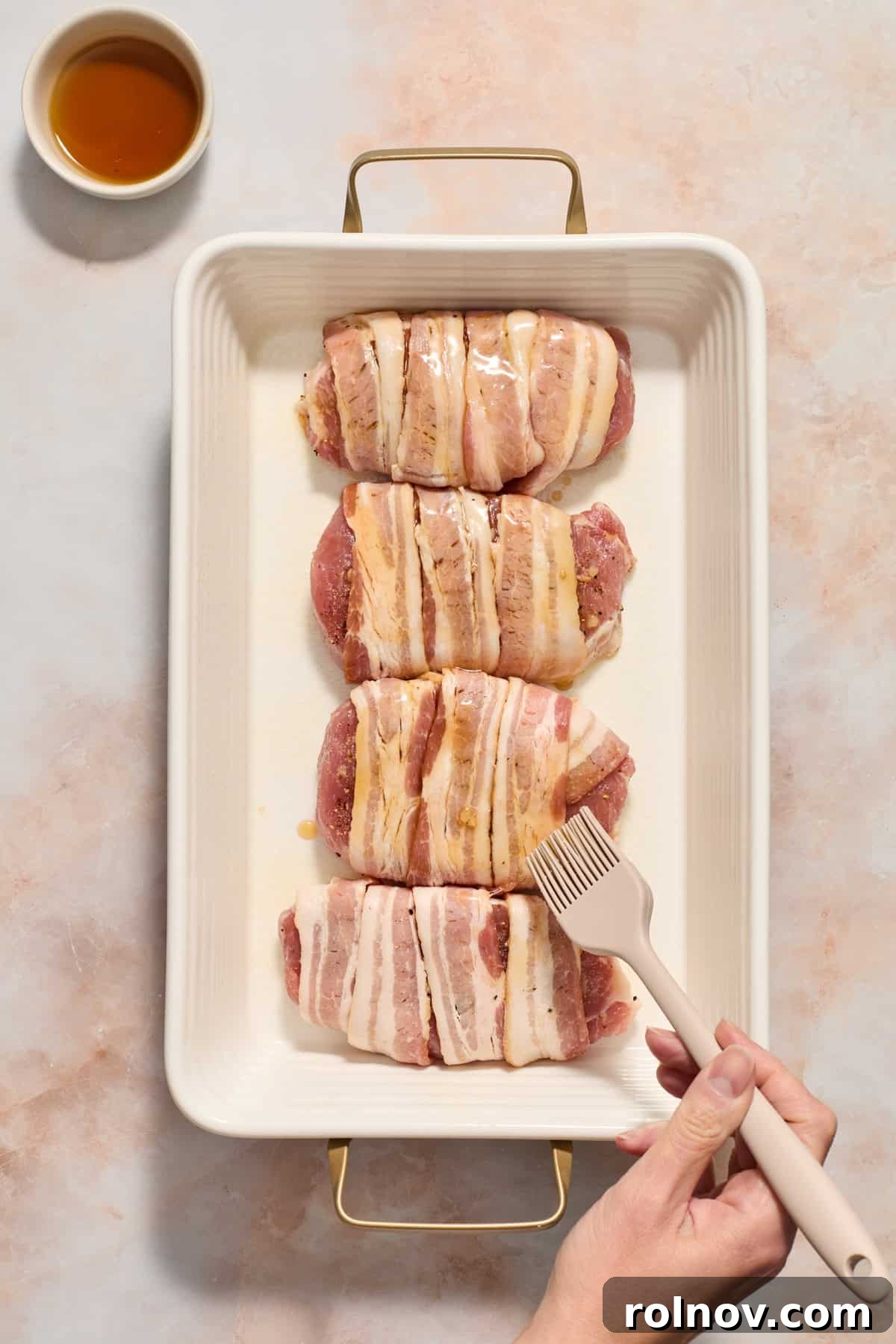 Sweet and Savory Maple Bacon Pork Chops 7 Maple syrup being brushed onto bacon-wrapped pork chops in a baking dish, with a small bowl of maple syrup next to the pan.