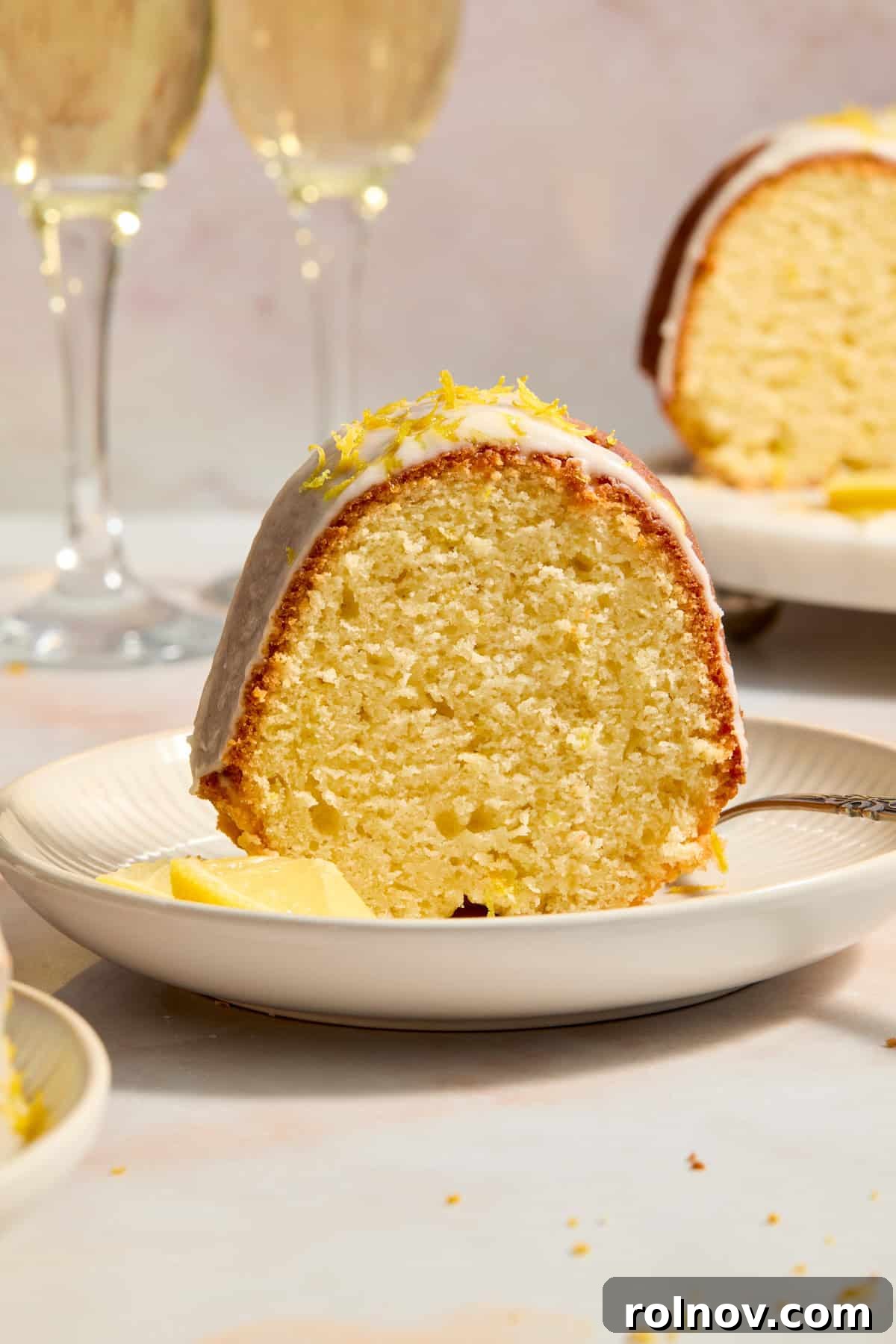 Effervescent Lemon Bundt Cake 10 A delectable slice of Champagne Lemon Bundt Cake served on a plate with fresh lemon slices, champagne glasses elegantly blurred in the background.