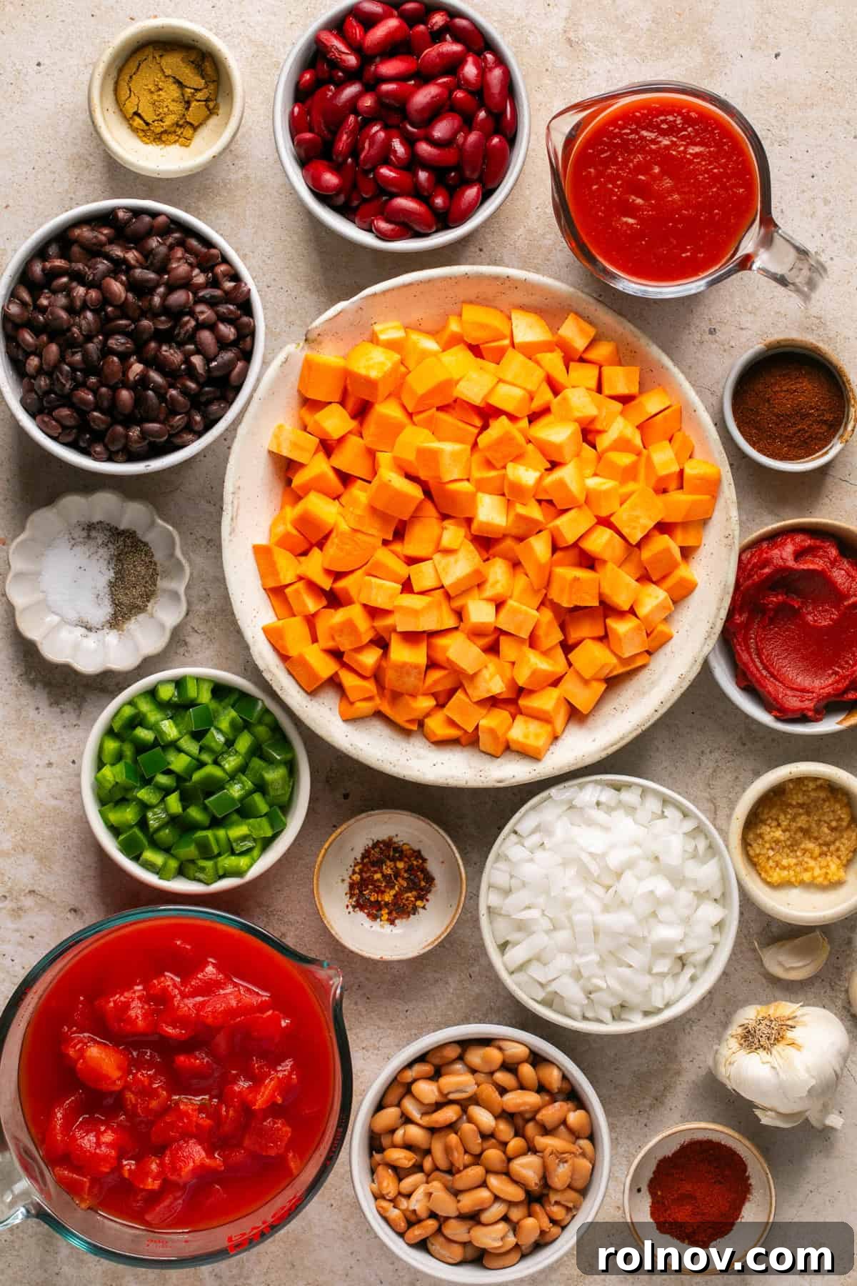 An array of fresh and canned ingredients for slow cooker sweet potato chili, including cubed sweet potatoes, various beans, canned tomatoes, spices, onion, and bell pepper, neatly arranged on a rustic surface.