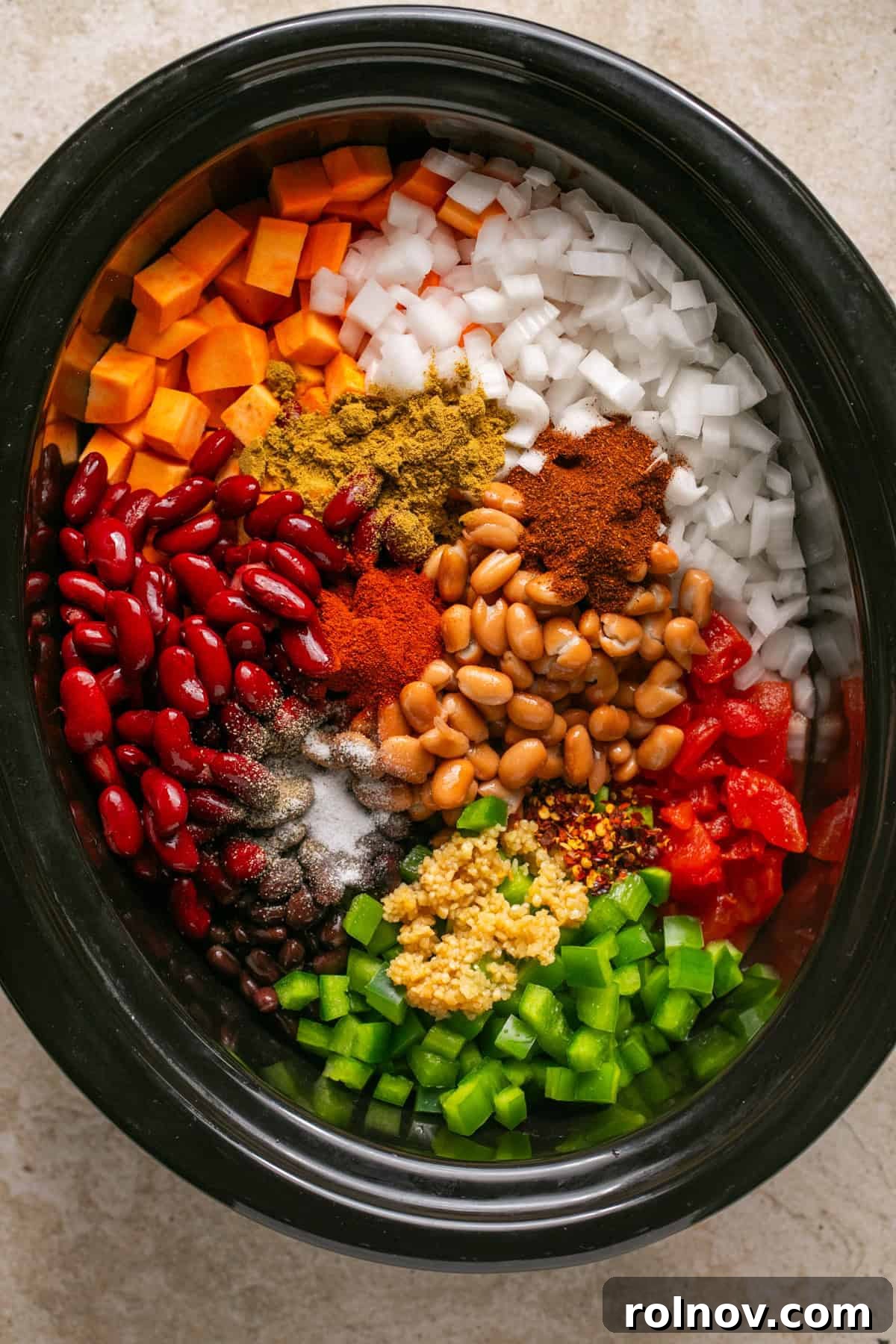 Diced sweet potatoes, various beans, green bell peppers, chopped onion, and spices arranged in a slow cooker, ready for stirring.