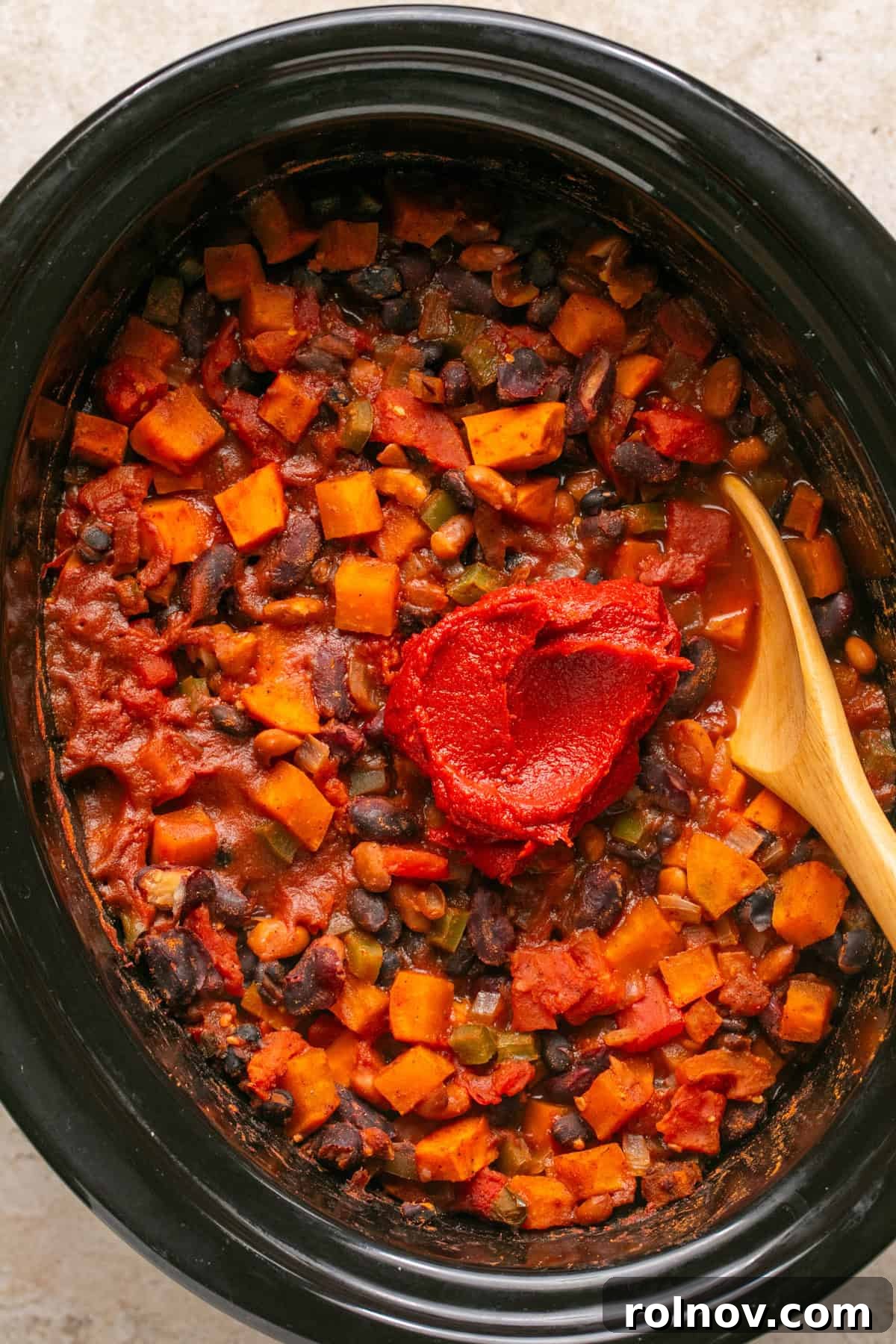 A dollop of rich tomato paste being added to the cooked chili in the crockpot with a wooden spoon, ready to be stirred in for thickening.