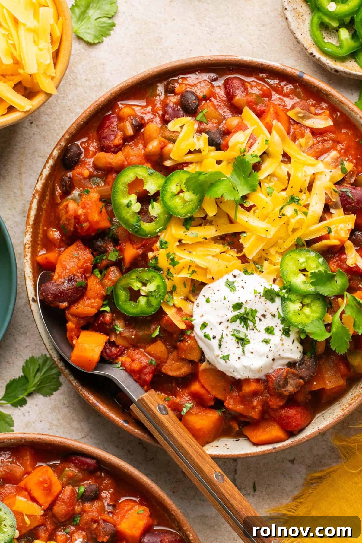 An overhead shot of a delightful bowl of slow cooker vegetarian chili, generously topped with shredded cheese, sour cream, and fresh jalapeño slices, with a spoon perfectly placed for scooping.