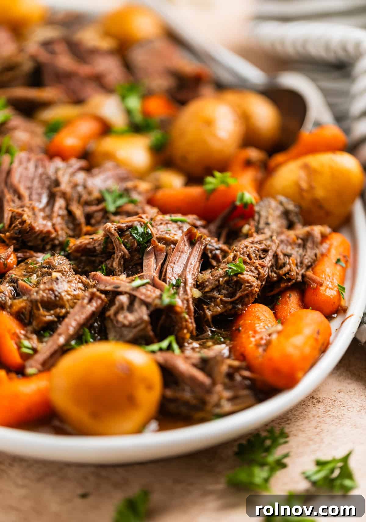 Slow Cooker Beef Pot Roast 2 Crockpot chuck roast on white serving platter with carrots, onion and petite potatoes and topped with chopped parsley.