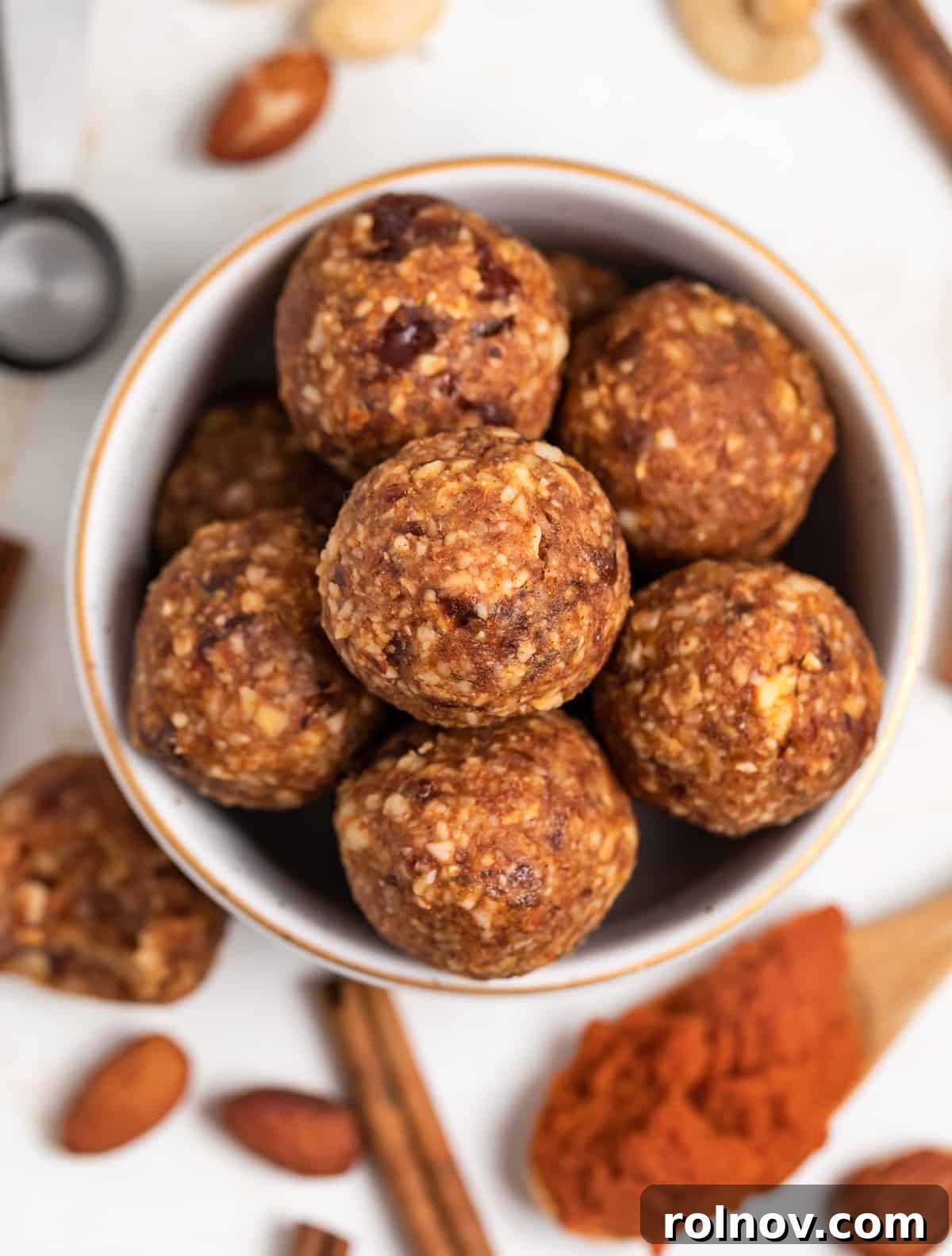 Pumpkin Spice Power Bites 2 A beautifully arranged bowl filled with golden pumpkin energy balls, surrounded by autumn accents like pecans, a cinnamon stick, a spoon of pumpkin puree, and raw cashews, embodying the rich flavors of fall.
