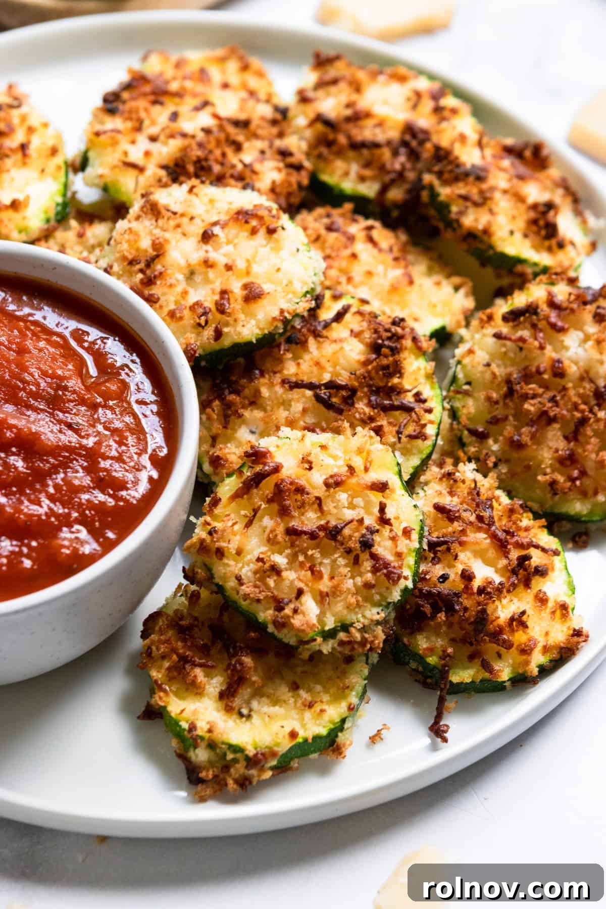Crispy Three Cheese Air Fryer Zucchini 2 Crispy Air Fried Zucchini rounds on a white plate with a side of marinara sauce for dipping.