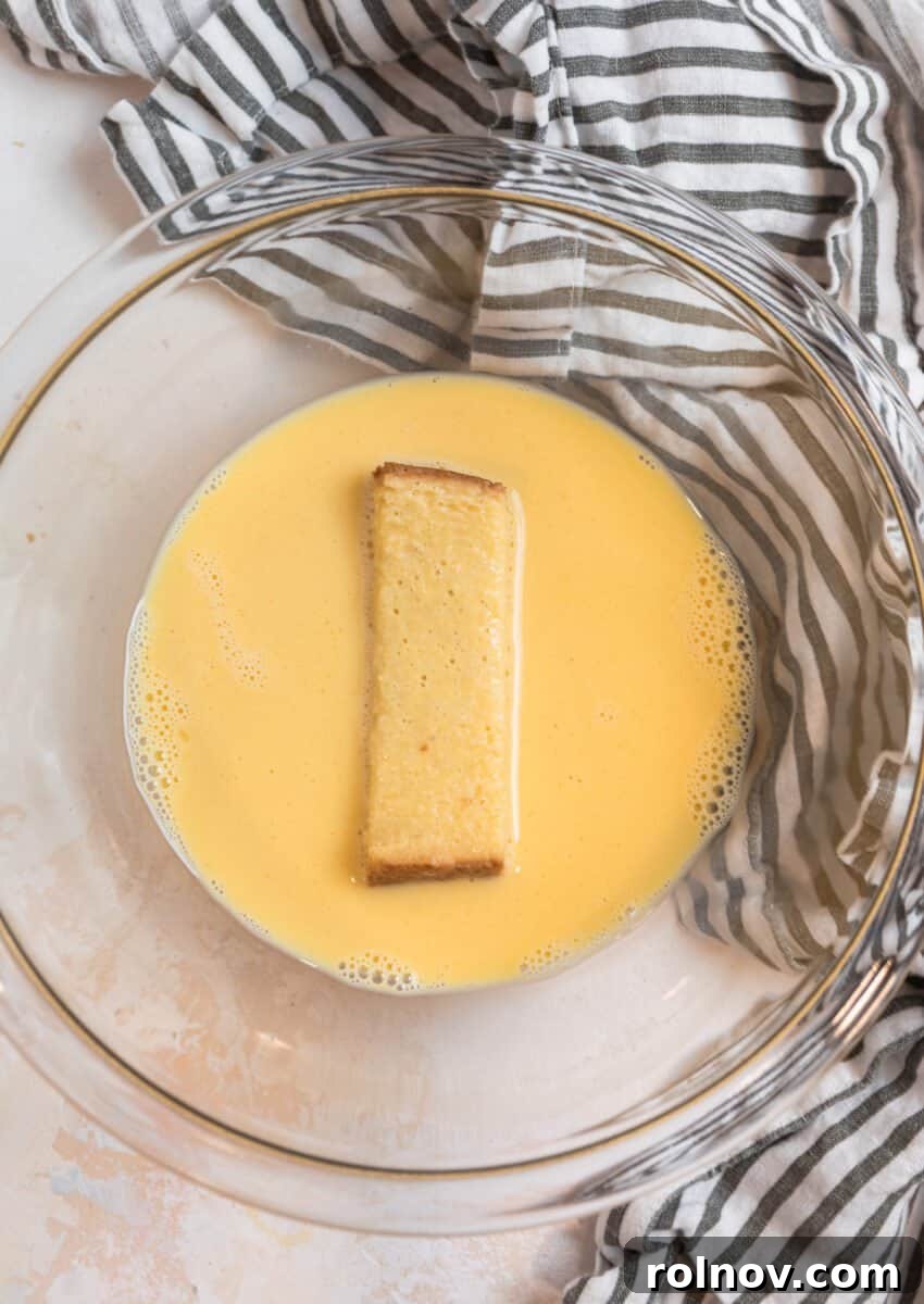 Crispy Air Fryer French Toast Sticks 5 A slice of bread being dipped into a bowl filled with the prepared egg mixture, showing the initial step of coating for French toast.